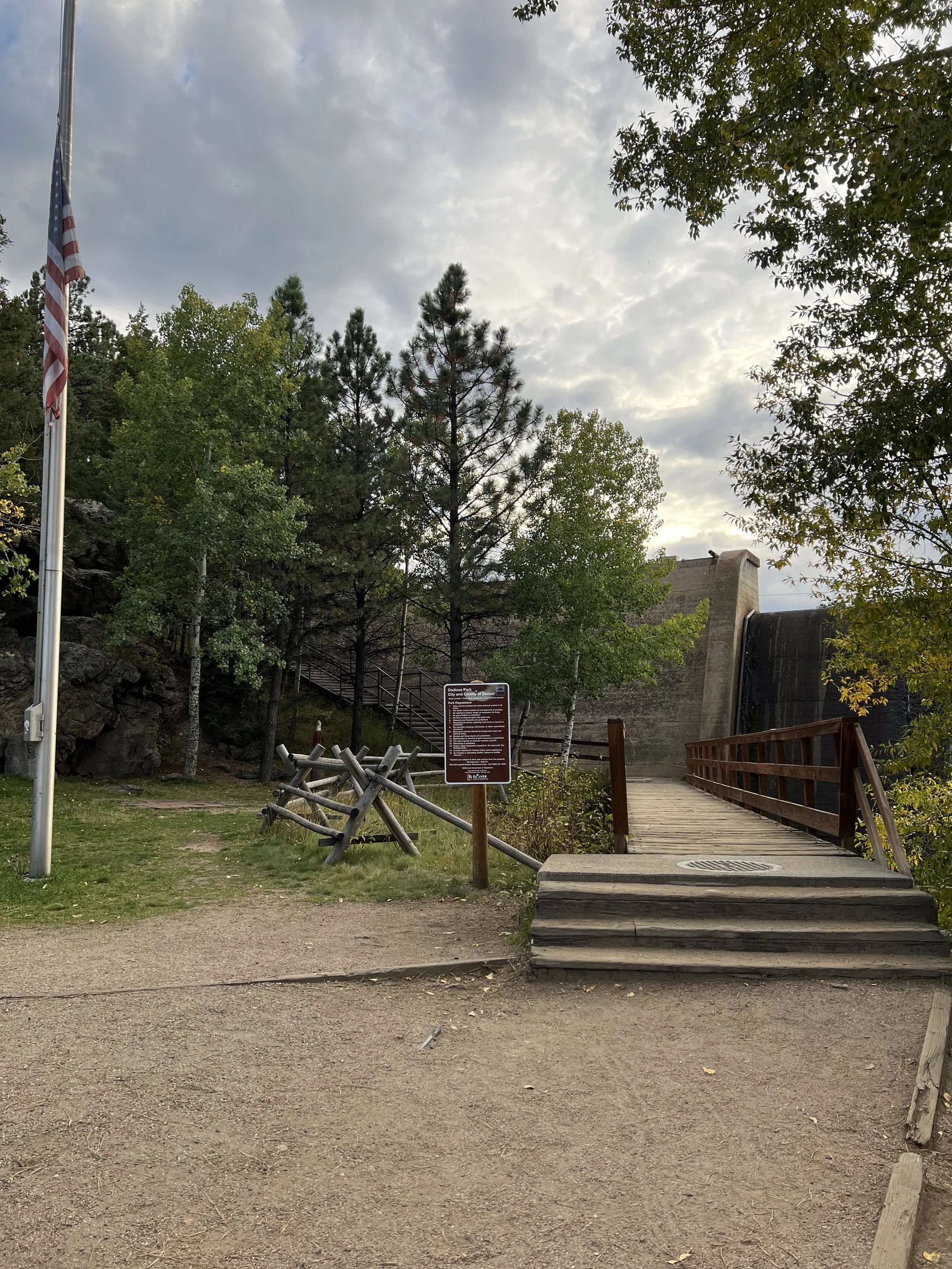 A park with a flagpole on the left, trees, a wooden bridge with stairs leading up to a dam structure, and an informational sign in the middle.
