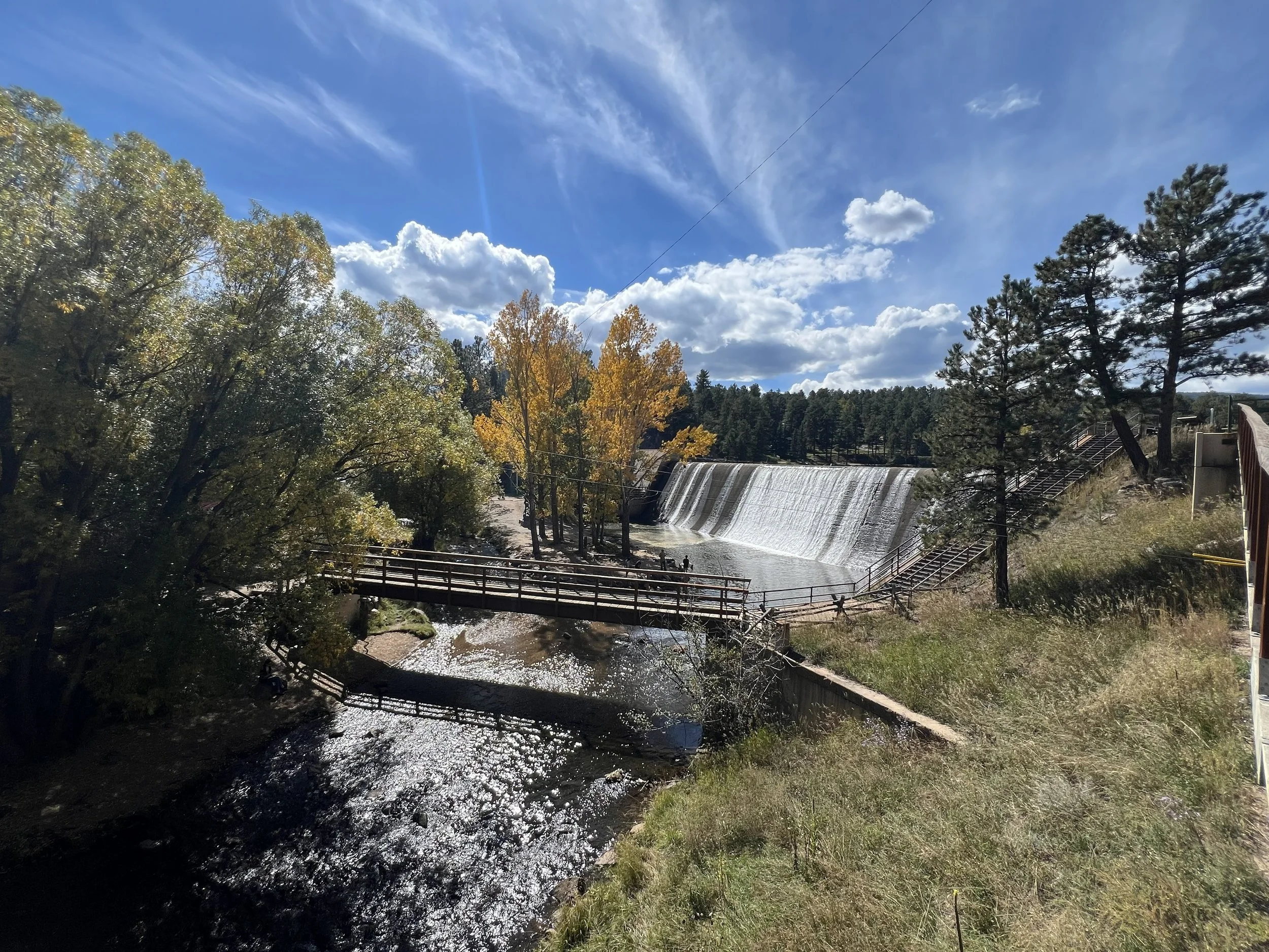 A scenic view of a waterfall with a curved dam, surrounded by trees with autumn-colored leaves, a blue sky with clouds, and a walking bridge over a river.