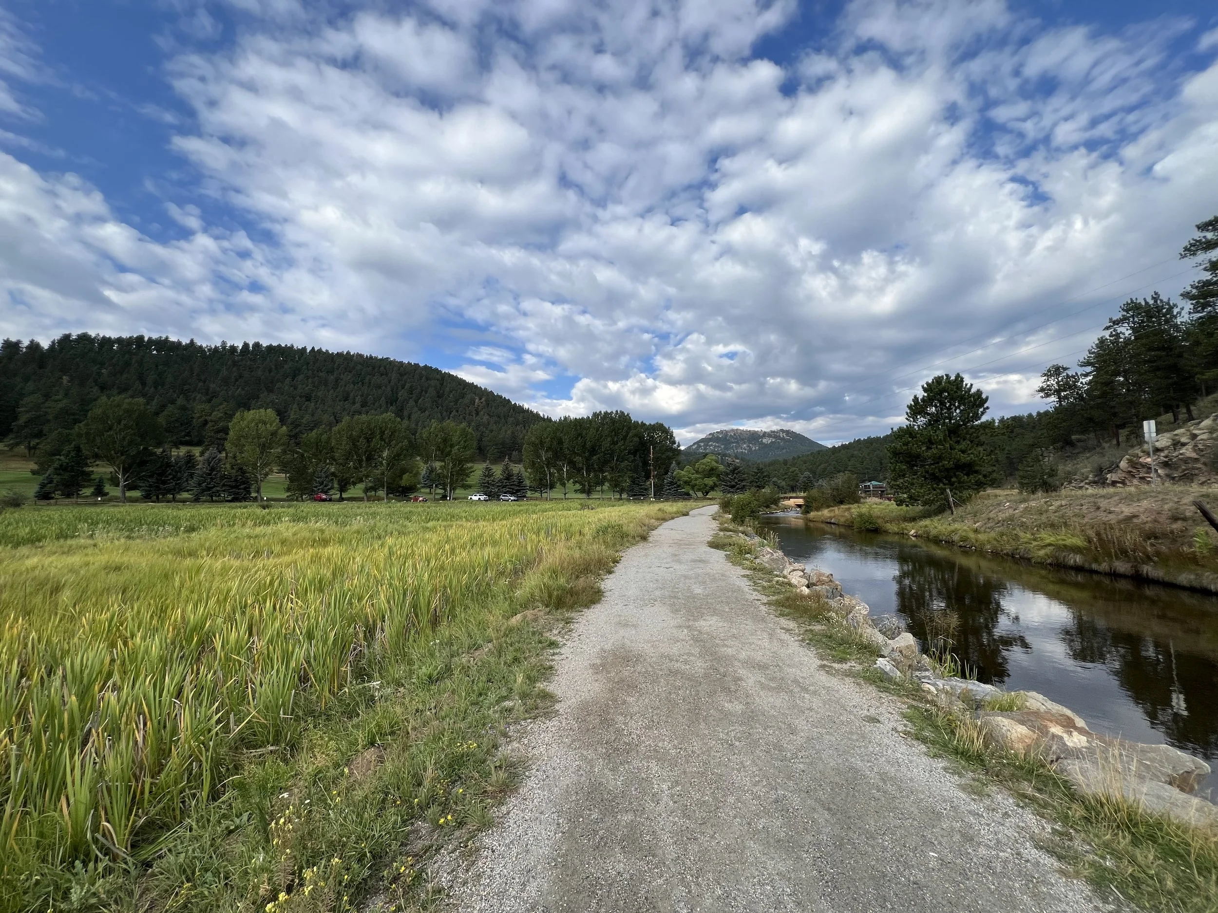 A gravel walking path runs beside a small river in a rural area with green fields, trees, and mountains under a partly cloudy sky.