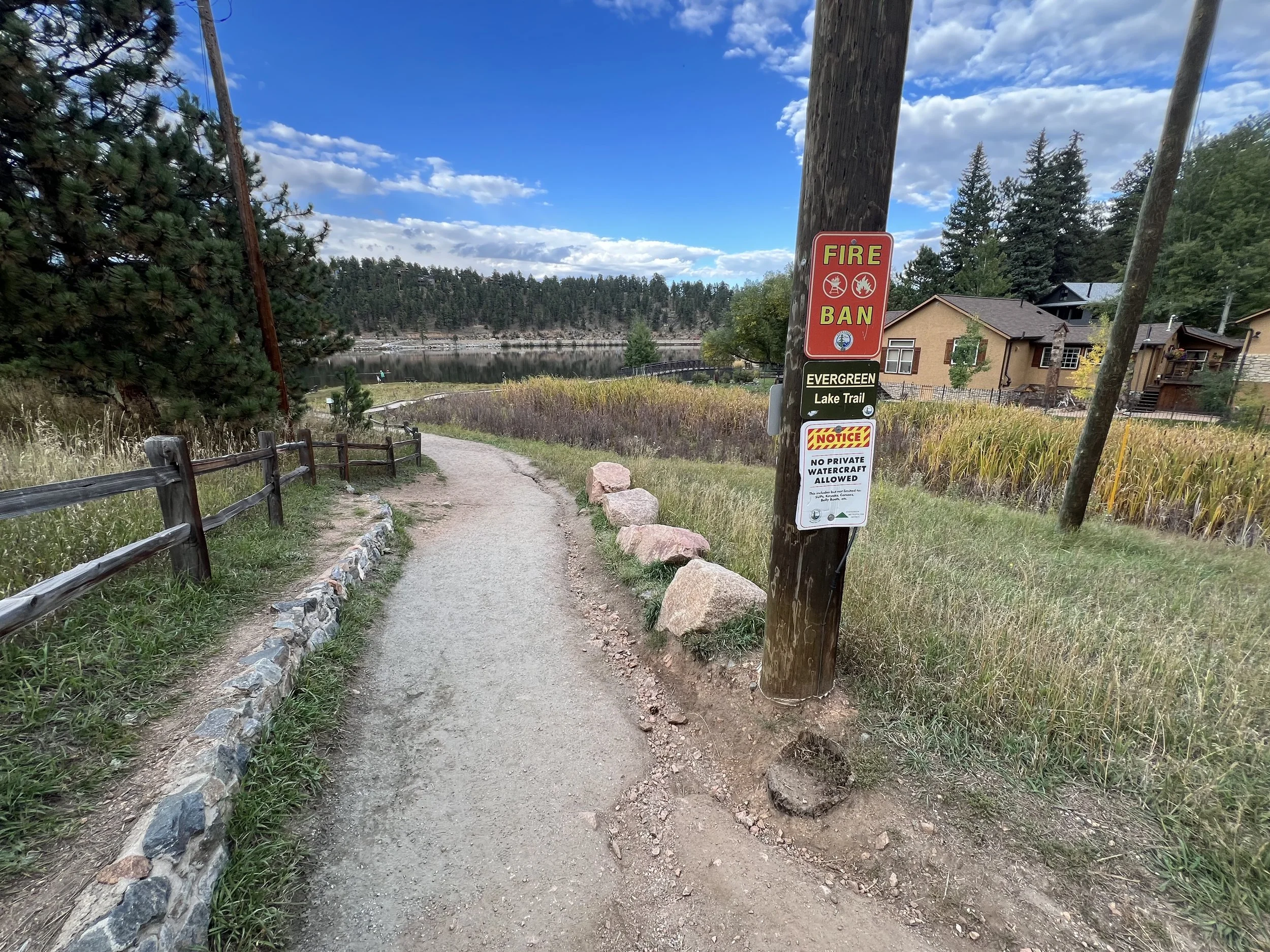 A dirt trail beside a lake with houses on the right and trees on the left. A sign on a utility pole near the trail reads 'Fire Ban' and 'No private watercraft allowed.' The sky is partly cloudy.
