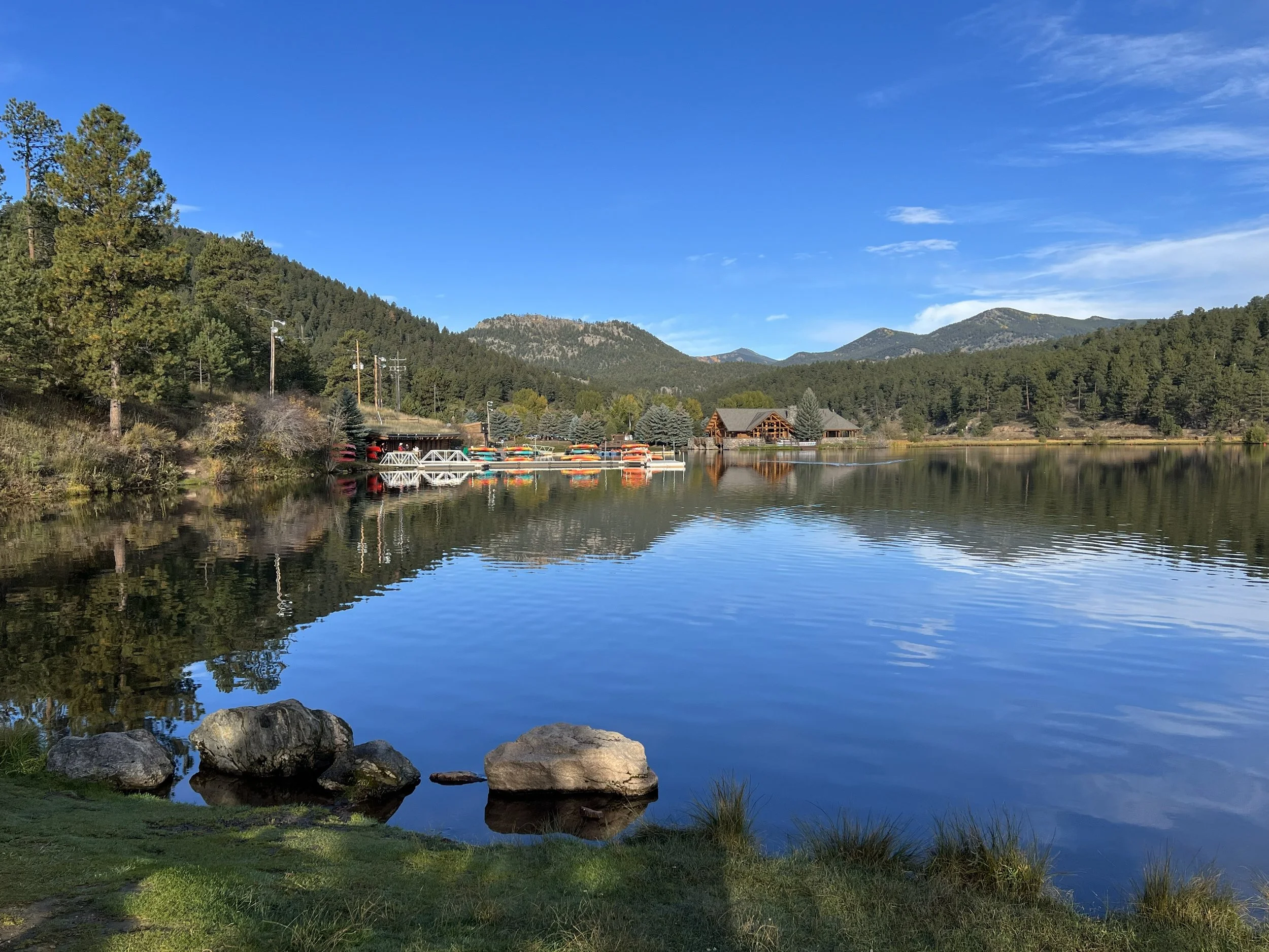 A serene lake with calm water reflecting the blue sky, green trees, and mountains in the background. There are rocks along the shoreline and a small dock with boats and a building nearby.