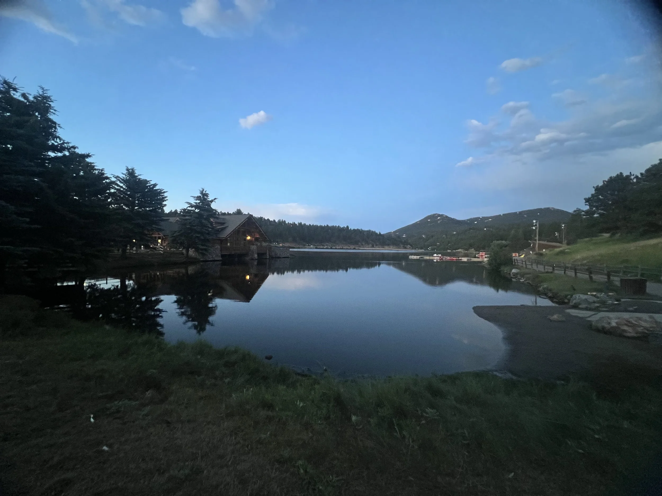 A serene lake surrounded by trees and hills, with a house on the left side and a mostly cloudy sky above.