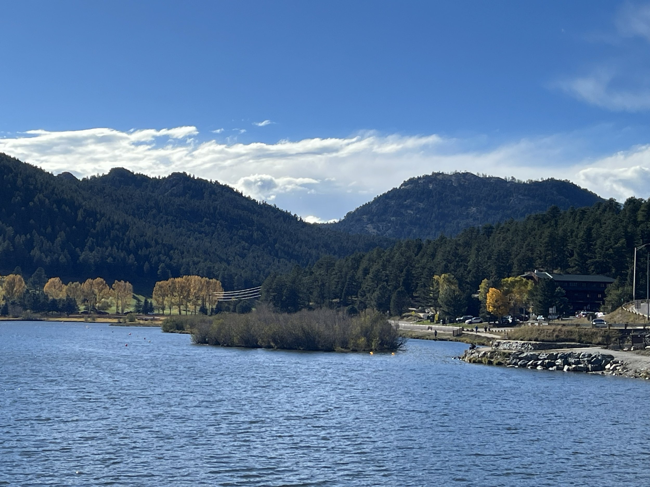 A calm lake surrounded by green mountains with a mix of evergreen and deciduous trees, some with fall foliage, under a partly cloudy blue sky.