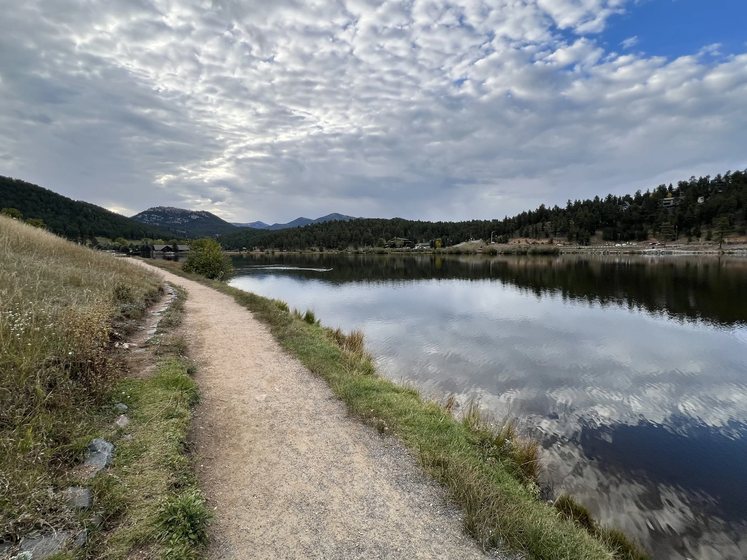 A dirt path runs alongside a calm river, surrounded by grassy hills and pine trees, with mountains in the background and partly cloudy sky overhead.