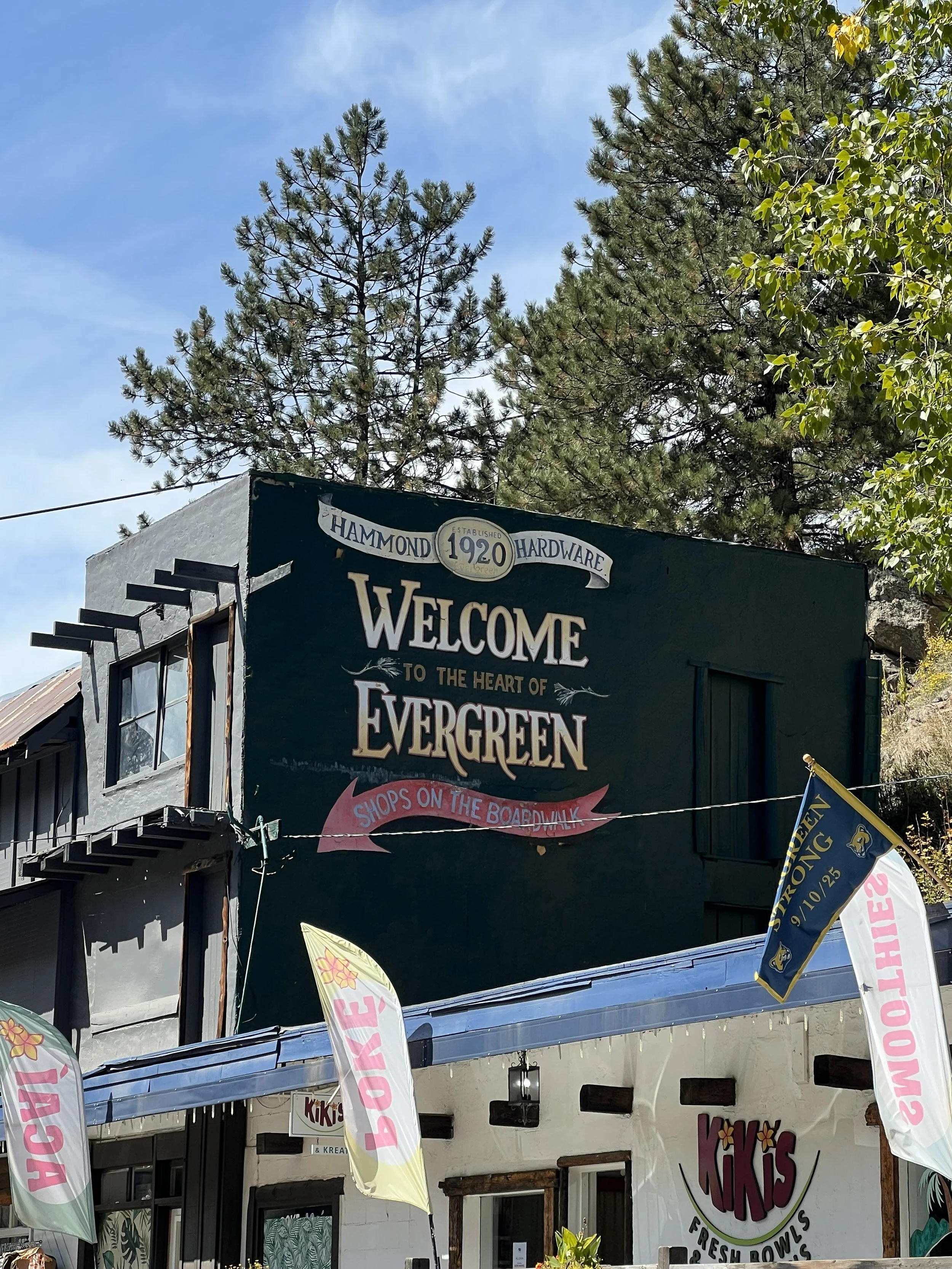 A sign on a building reads 'Welcome to the heart of Evergreen,' with smaller text mentioning shops on the boardwalk. The sign indicates the establishment was founded in 1920 and sells hardware. Nearby are flags, including one with 'Strong' and a date