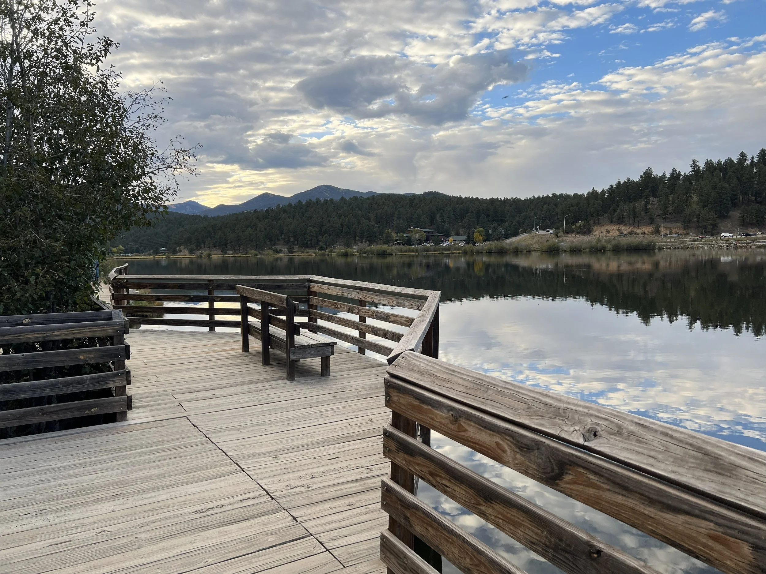 A wooden pier extending over a calm lake with a mountain range in the background, surrounded by trees and a partly cloudy sky reflected in the water.