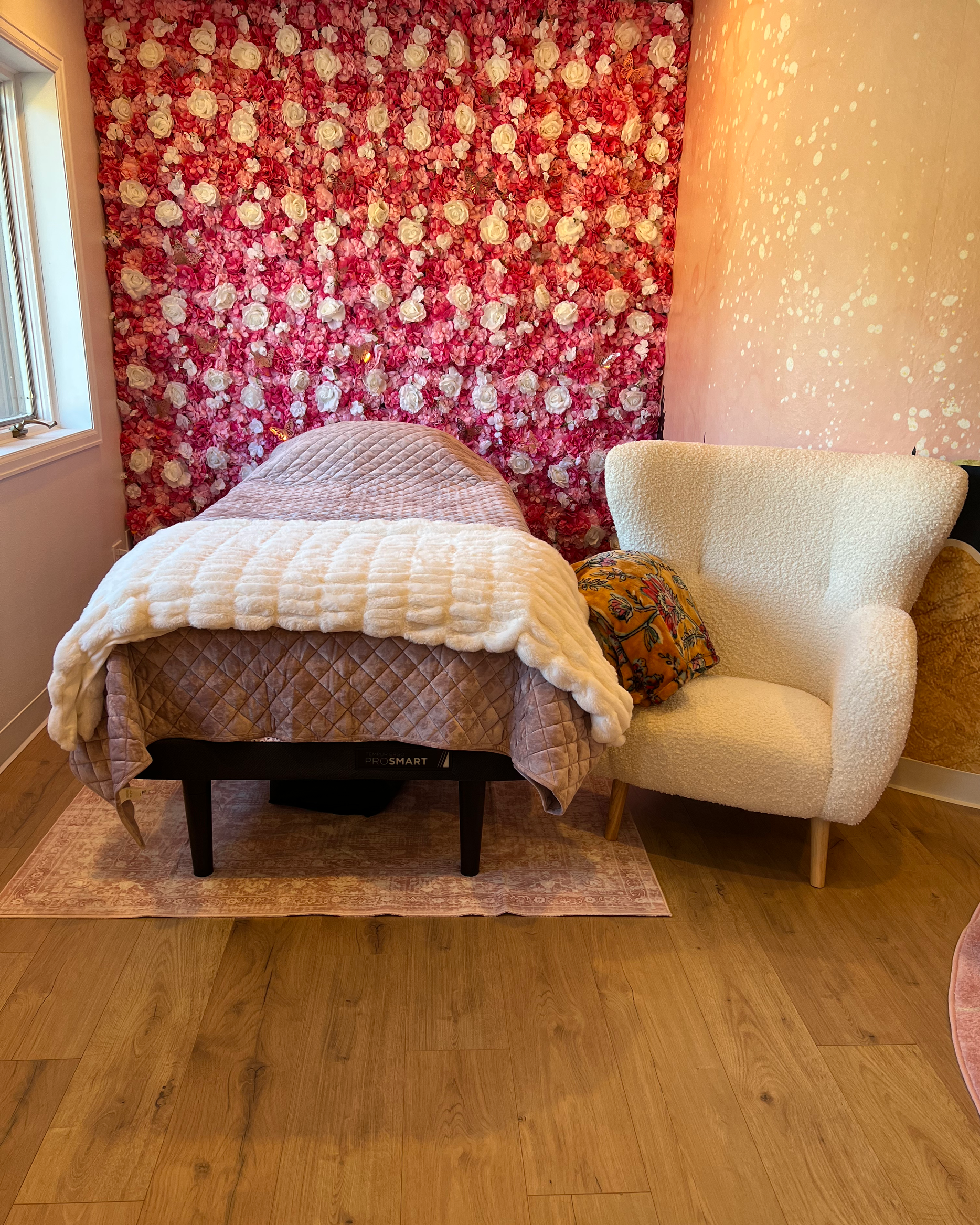 A room with a bed covered with a quilted beige bedspread and a plush white blanket, positioned in front of a floral pink and white flower wall. To the right of the bed is a cream-colored armchair with a pillow, partially overlapping the bed. There's 