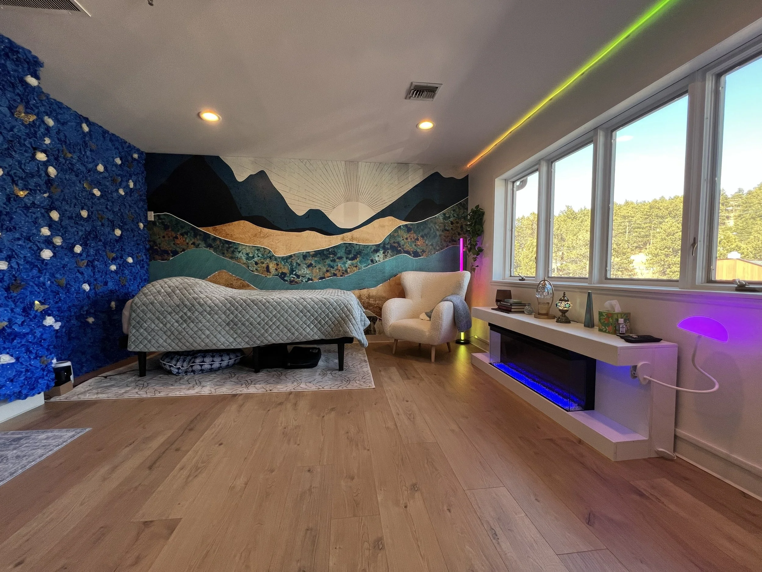 Living room with a mountain mural, blue floral accent wall, white armchair, long couch covered with a quilt, large window showing outdoor trees, and a modern fireplace with decorative items on the mantel. Colorful LED lights add ambiance.