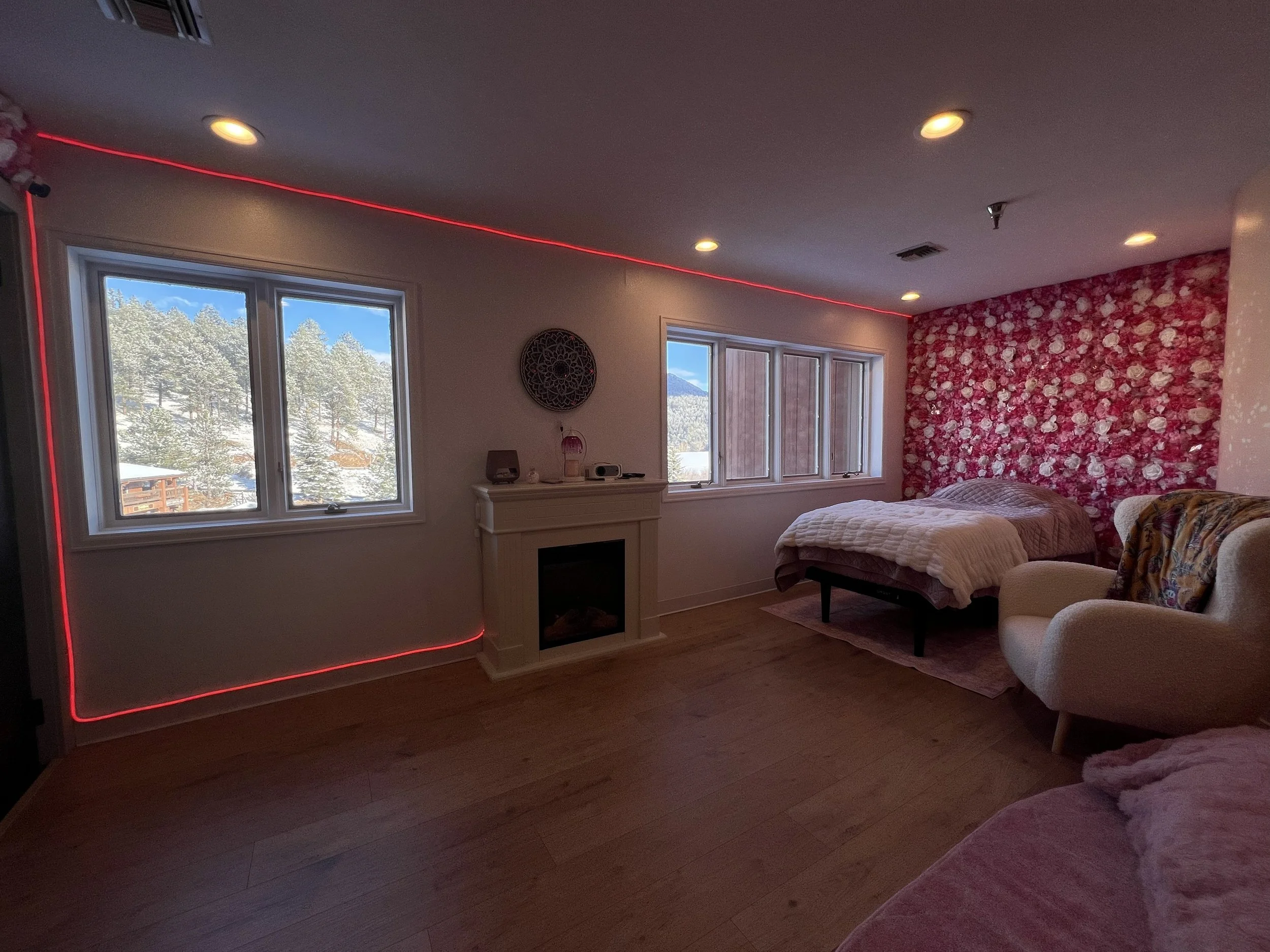 A bedroom with large windows showing a snowy landscape and mountains outside. The room has a pink floral accent wall behind the bed, which has a white quilt. There's a beige armchair with a colorful throw in the corner, and a fireplace mantel with sm