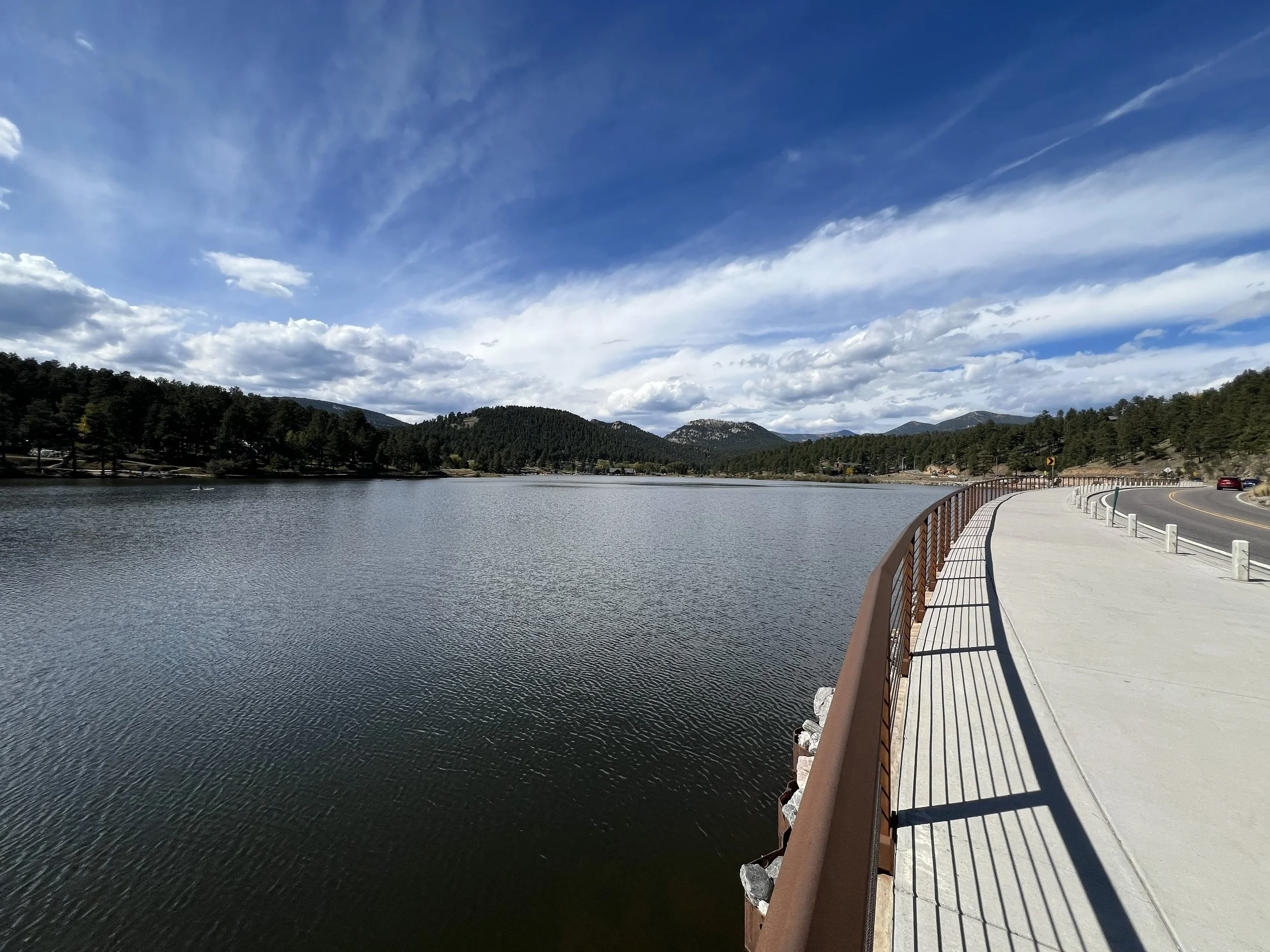 A scenic view of a lake with a paved walkway and a rusted metal railing on the right, trees on the far side of the lake, mountains in the distance, and a partly cloudy sky overhead.