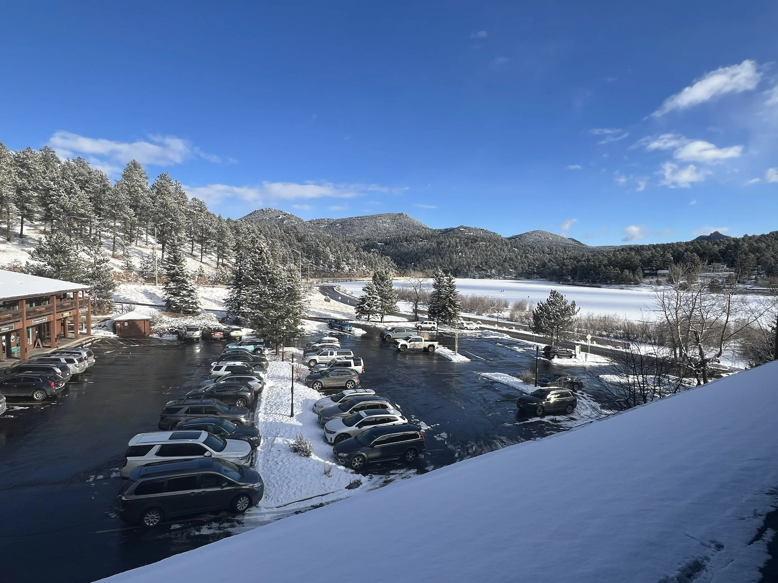 Snow-covered parking lot with parked cars, a building on the left, snow-covered trees, and rolling mountains in the background under a partly cloudy blue sky.