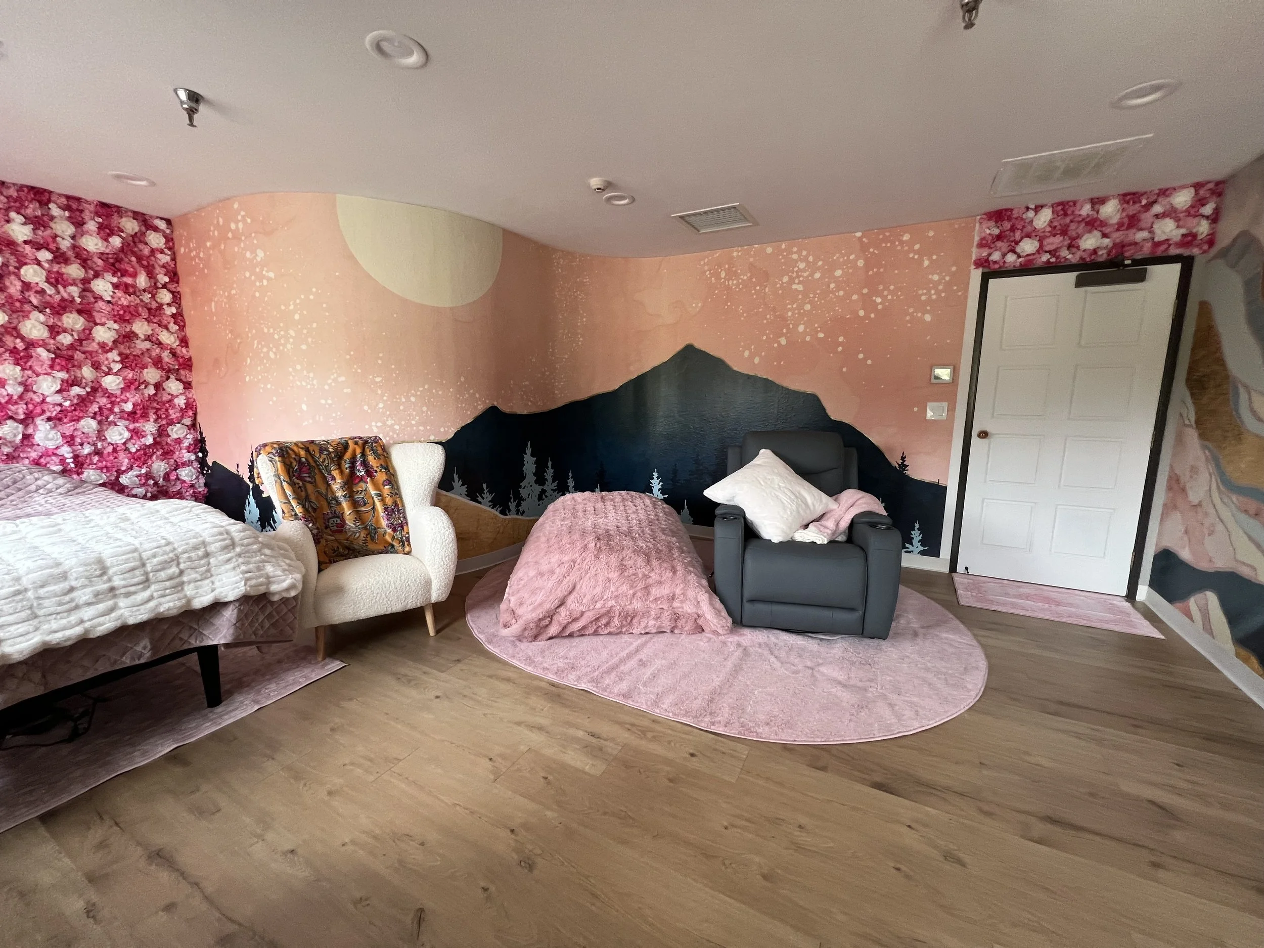Living room with pink and black mountain mural, pink circle rug, pink blanket, white armchair with floral blanket, black armchair with white pillow, bed with beige bedding, floral wall in background and a white door.