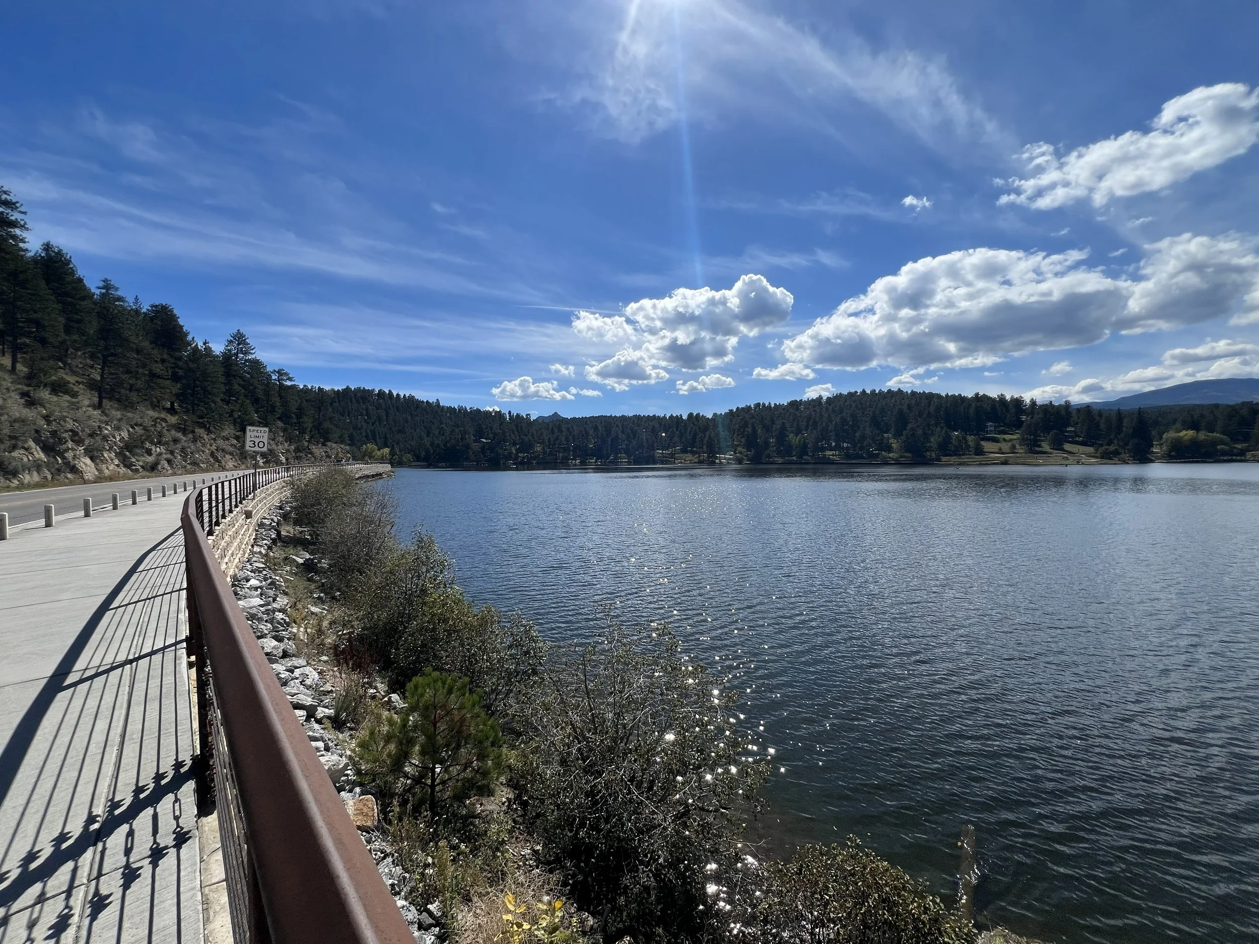 A scenic view of a lake with a concrete pathway and metal railing on the left, surrounded by forests and mountains under a partly cloudy blue sky with the sun shining.