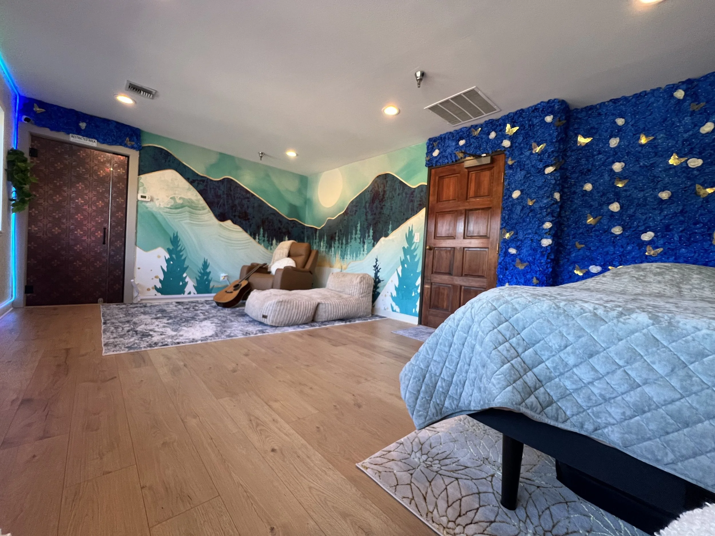 A room with a mountain and forest mural on the wall, a recliner chair, a bean bag, an acoustic guitar, a bed with a quilt, and decorated wall panels with blue and gold butterflies.
