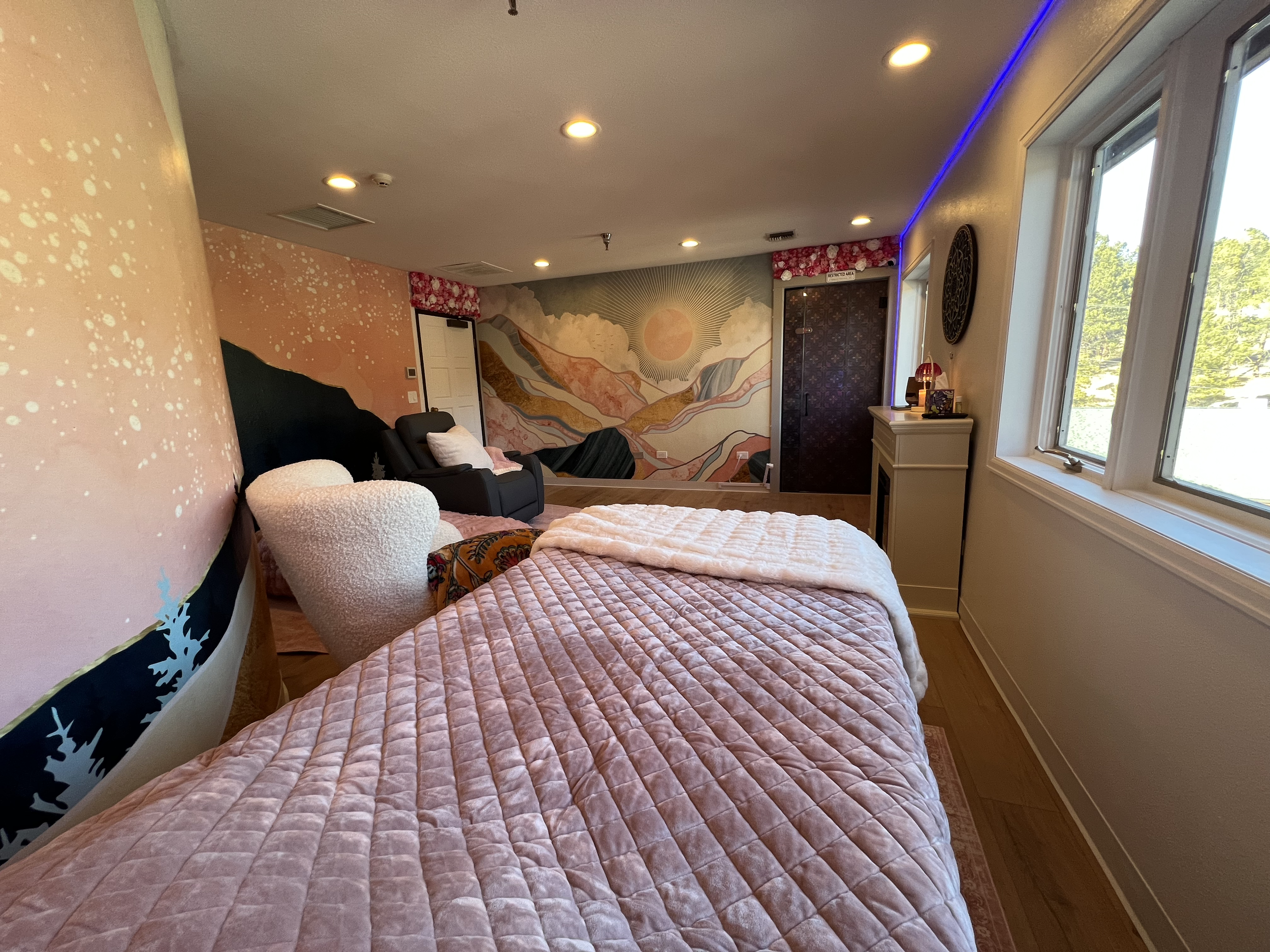 A bedroom with a pink quilted bedspread, a white plush armchair, and a dark grey recliner with pillows. The wall behind has a colorful mountain landscape mural, and there are windows on the right side showing green trees outside. The ceiling has rece
