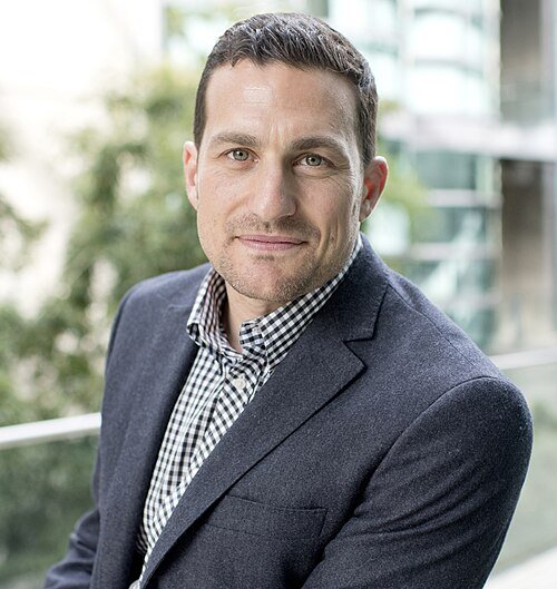 A man with short dark hair and light skin, wearing a dark blazer and checked shirt, standing outdoors with a blurred cityscape background.