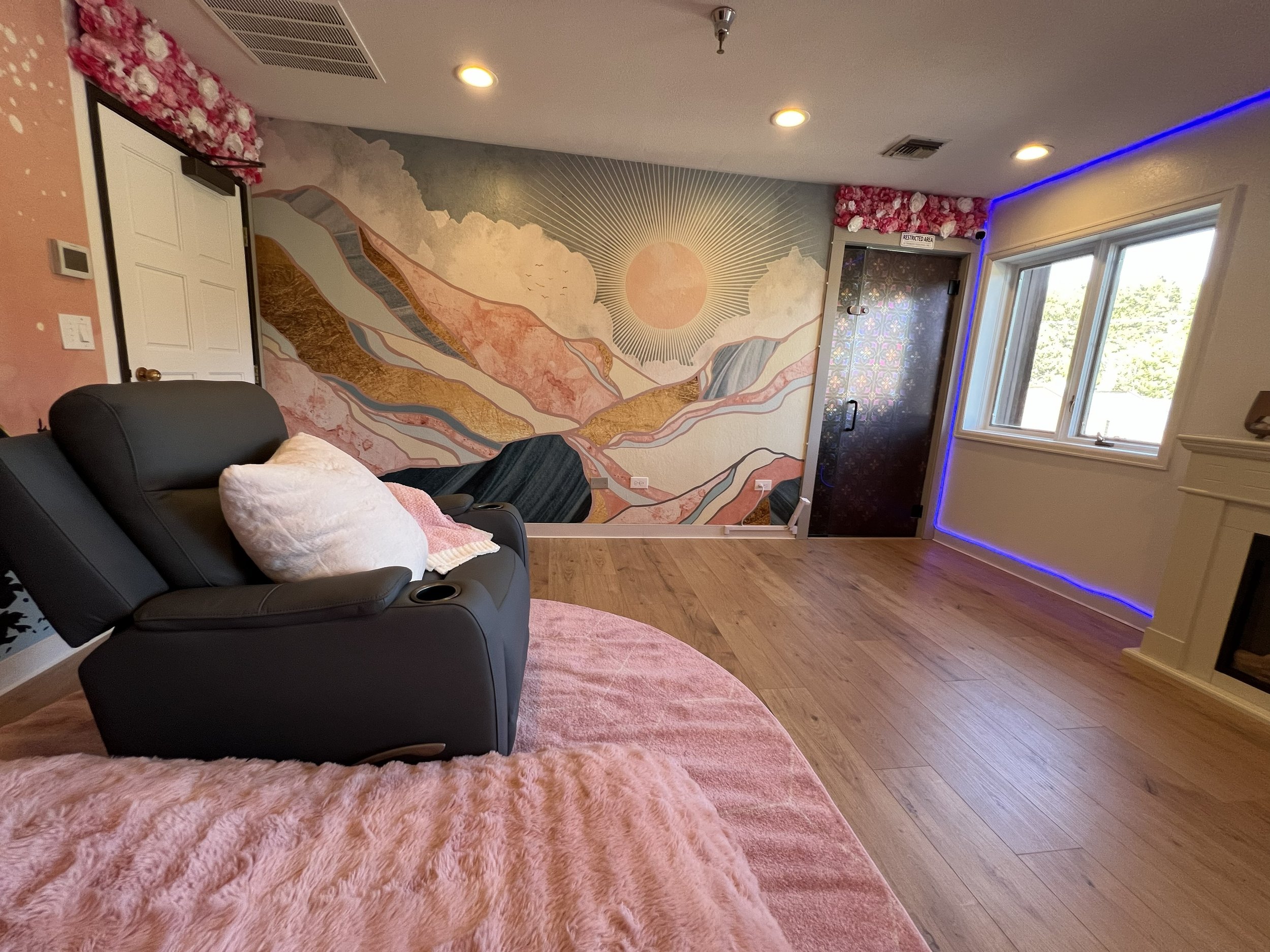 Room with a mural of mountains and the sun on the wall, black massage chair with a white pillow, pink blanket, pink furry rug, wooden floor, large window, and LED strip lighting on the ceiling and wall.