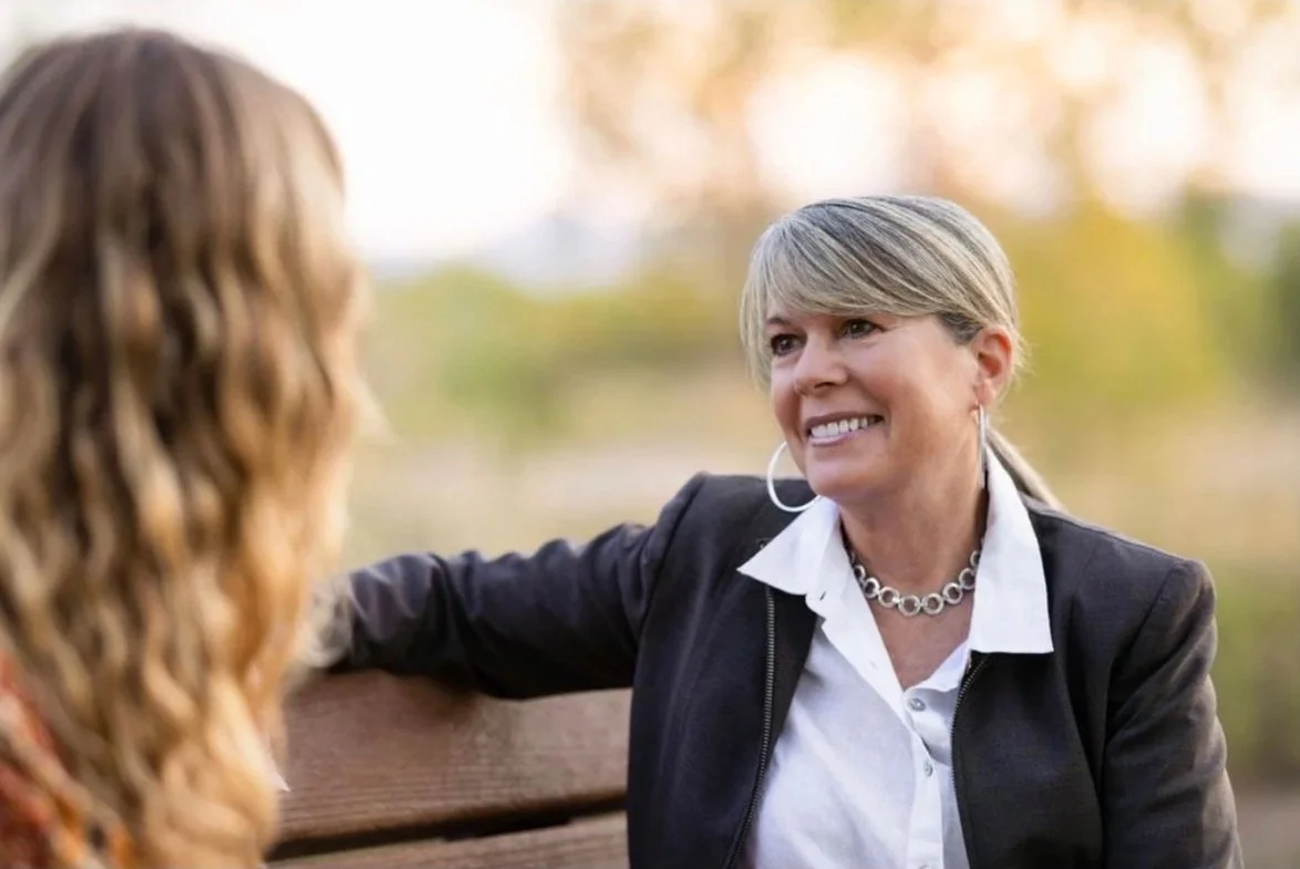 Two women having a conversation outdoors, one with blonde hair and the other with light brown, sitting on a park bench with autumn trees in the background