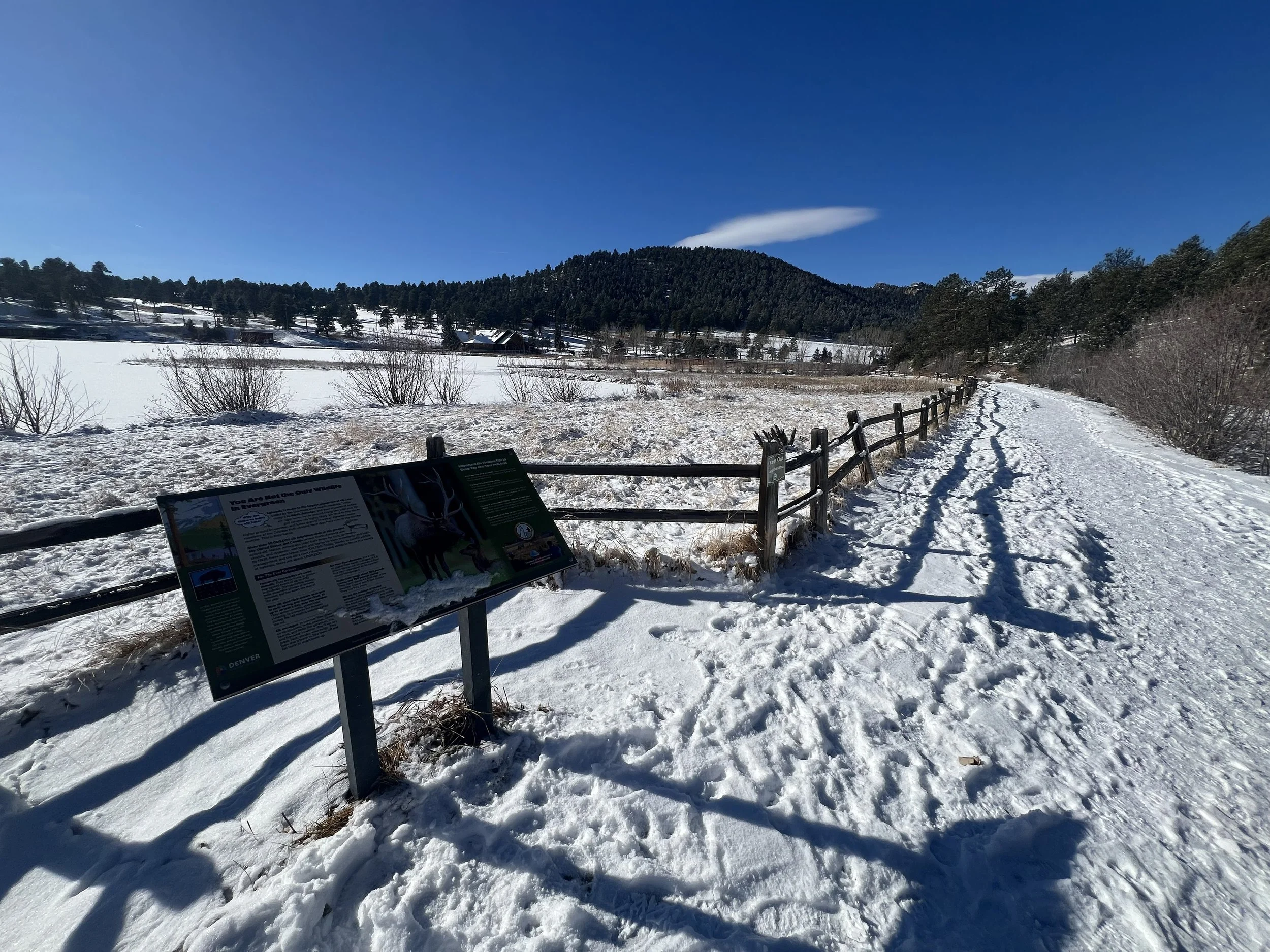 Snow-covered trail with wooden fence and informational sign, field, trees, hills, and clear blue sky in the background.