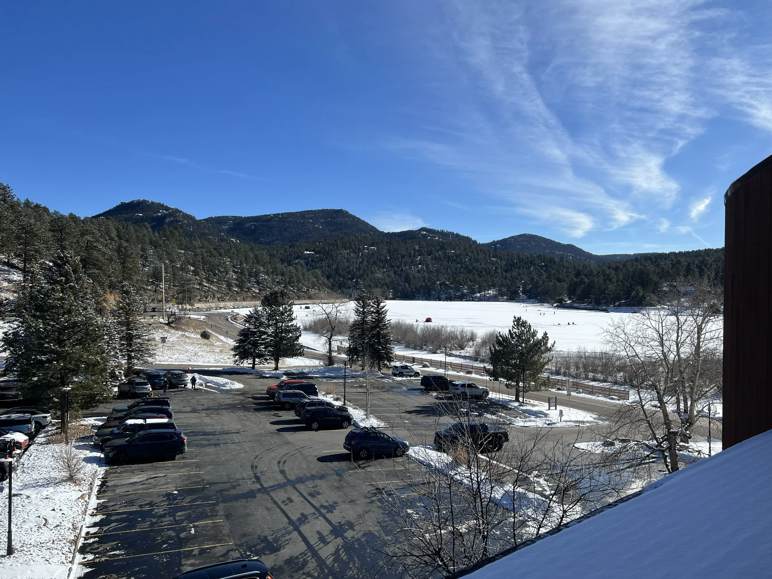 Snow-covered parking lot with cars, trees, and distant mountains under a clear blue sky.
