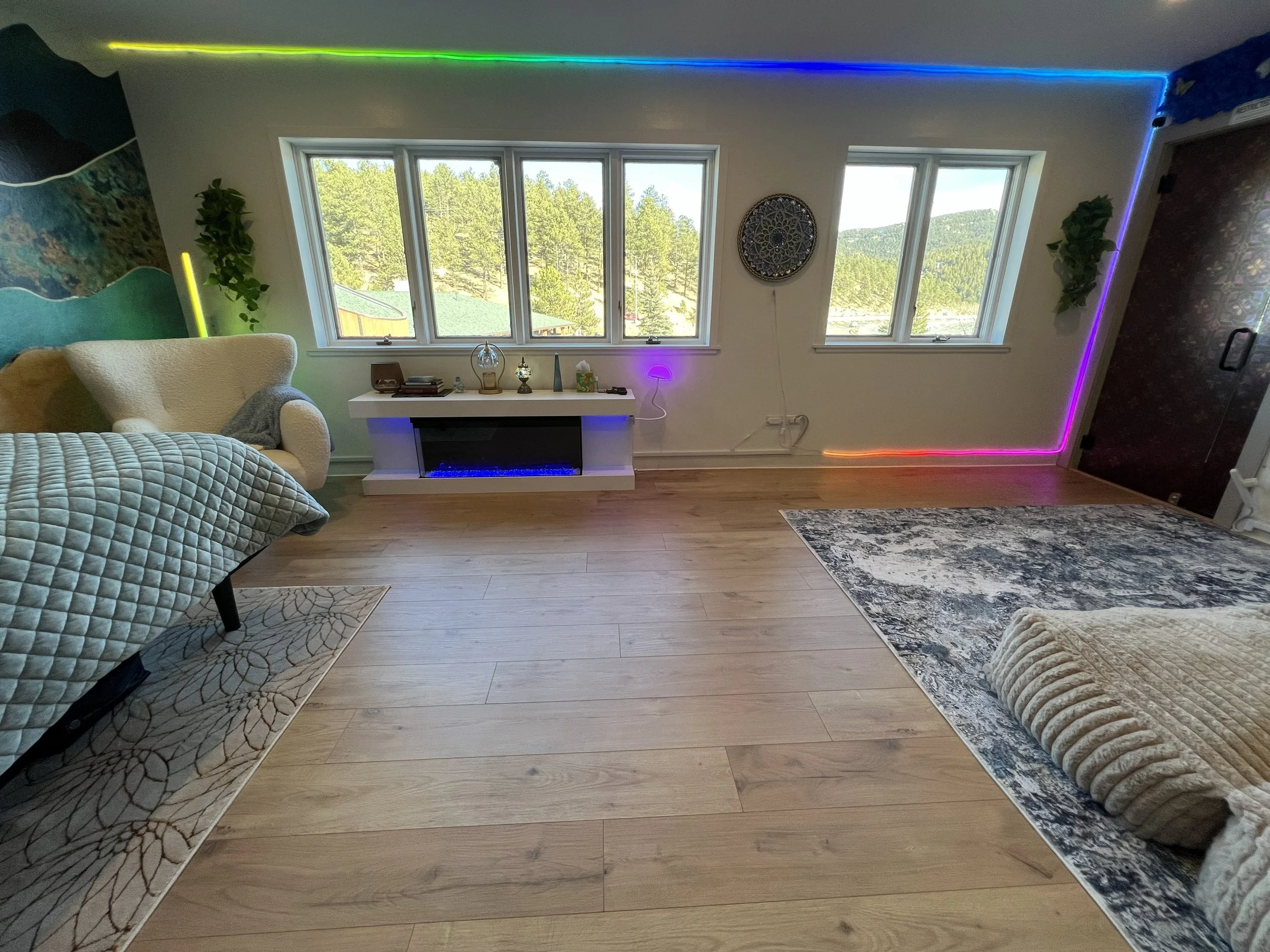 Living room with wooden floor, a window with a view of green trees and mountains, a beige armchair, a patterned blanket, a modern fireplace with blue light, decorative items on the mantel, a rug, and LED strip lights in multiple colors along the ceil