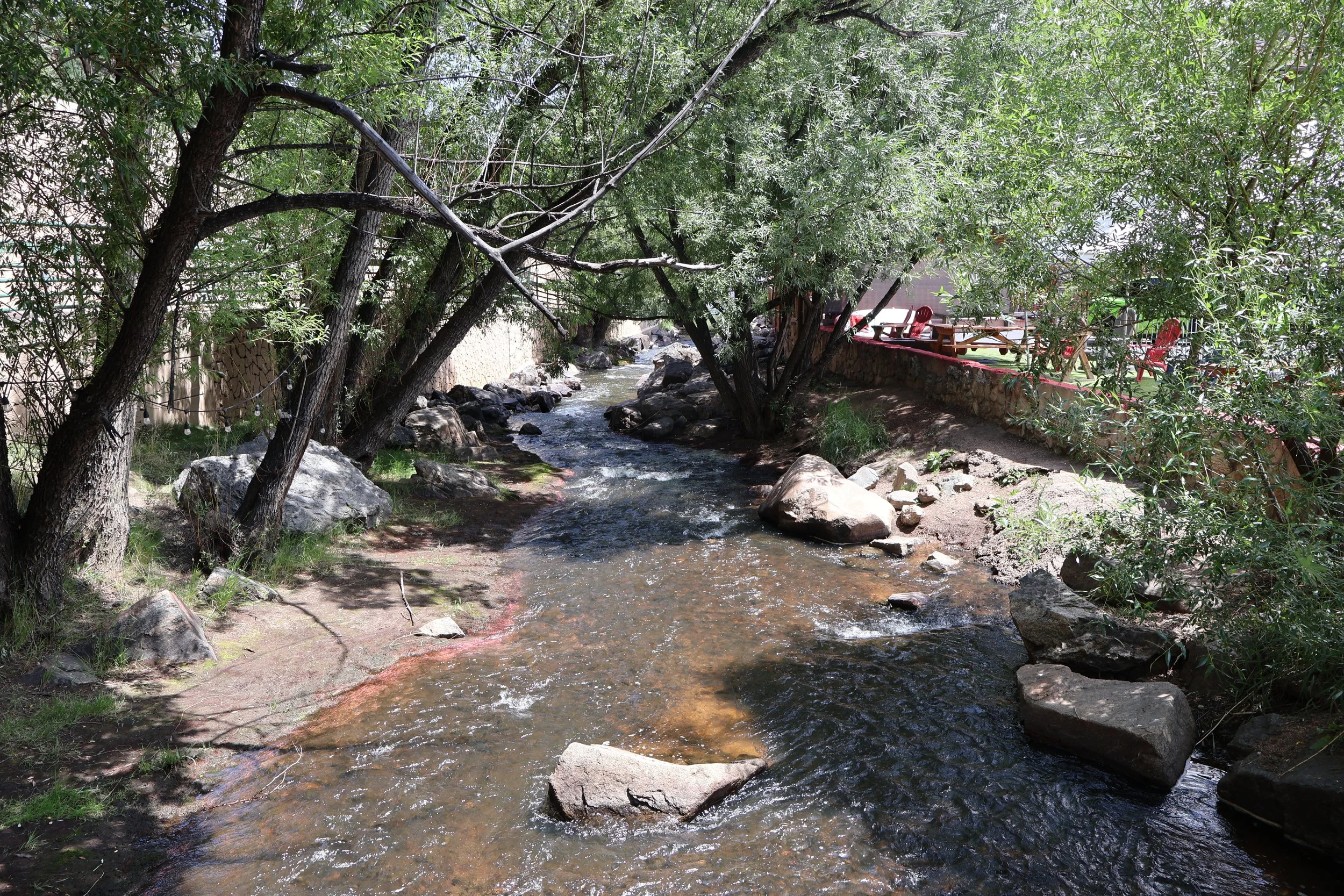 A small stream flowing through a shaded area with trees on both sides and some outdoor seating in the background.
