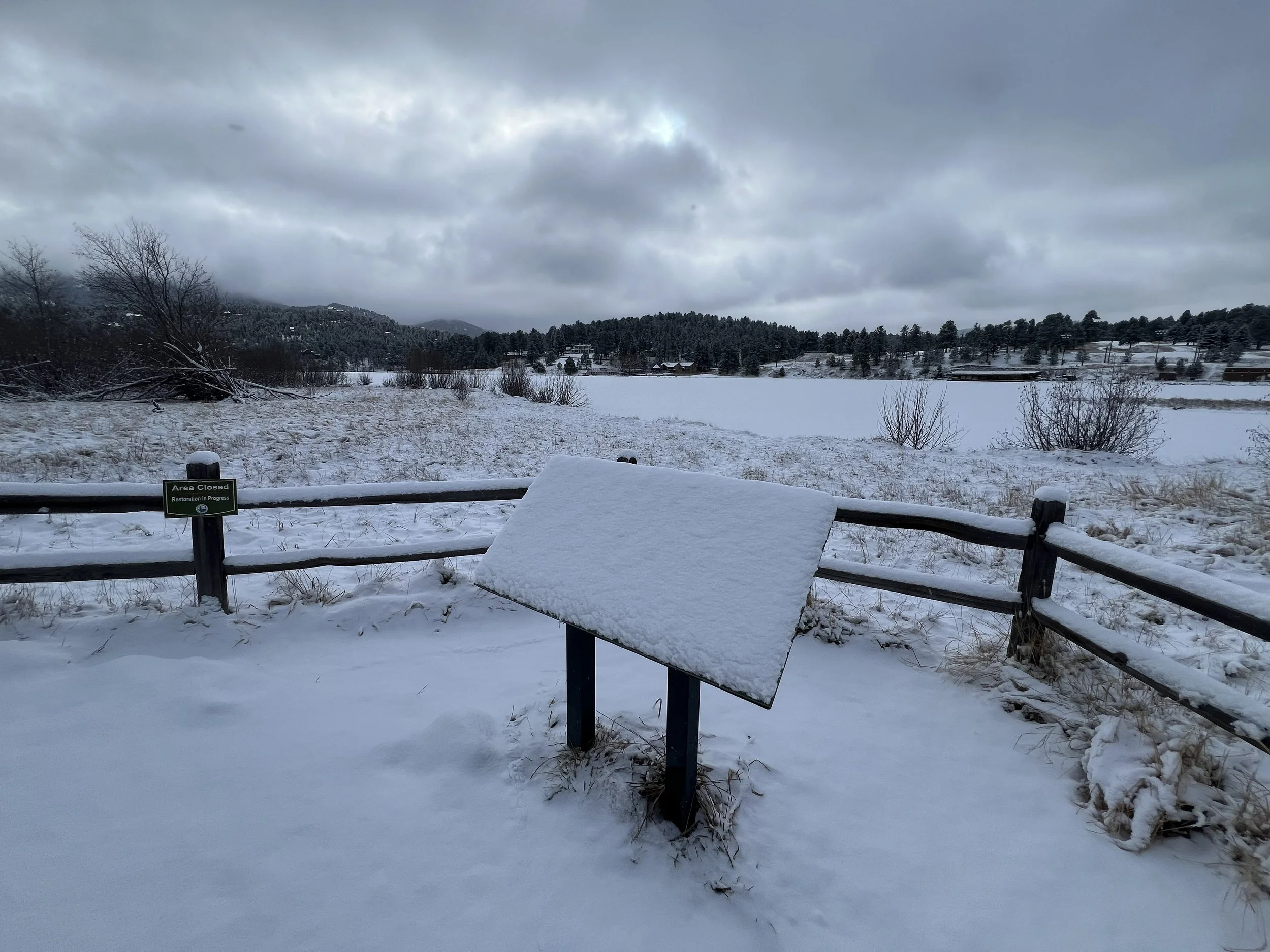 Snow-covered landscape featuring a frozen lake or river, a wooden fence with a sign that reads 'Area Closed, Restoration in Progress,' and mountains in the background under a cloudy sky.