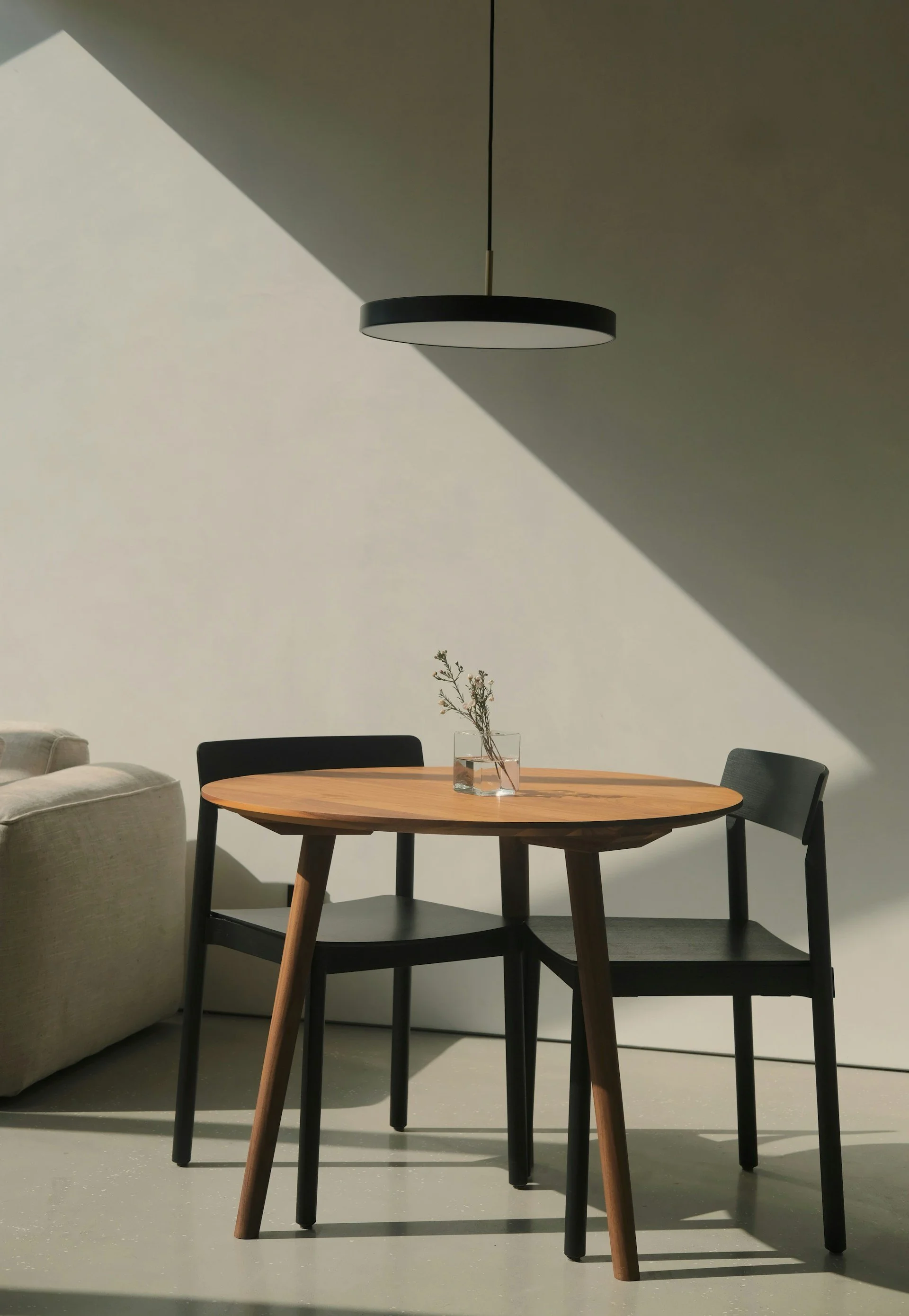 Mesa de madera redonda con dos sillas negras, una con respaldo curvado, en una habitación con luz natural y sombra, con una lámpara circular negra en el techo y una vasija con pequeñas ramas secas.