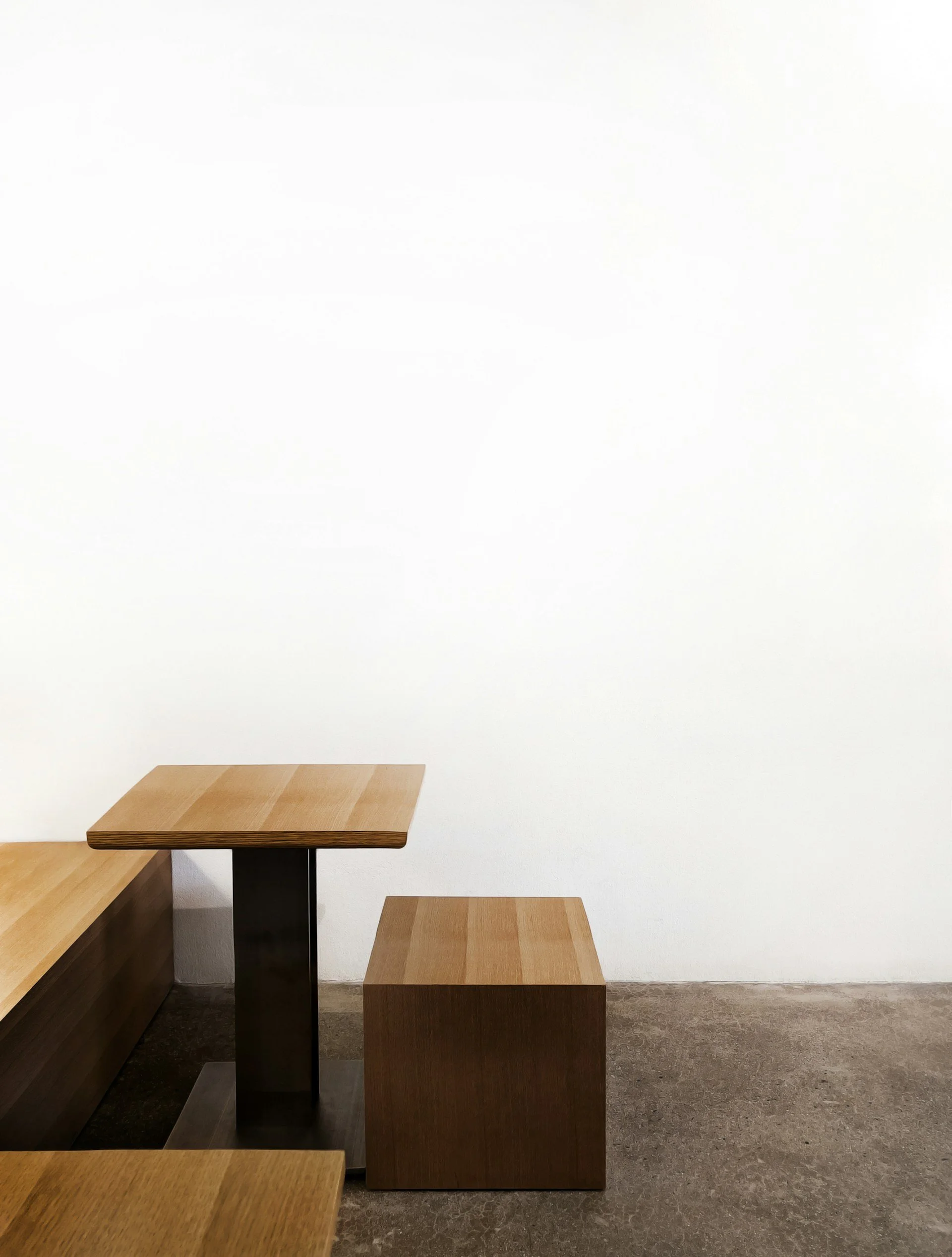 Muebles de madera, una mesa pequeña sobre un soporte de metal y un cubo de madera, en un espacio minimalista con pared blanca y suelo de concreto.