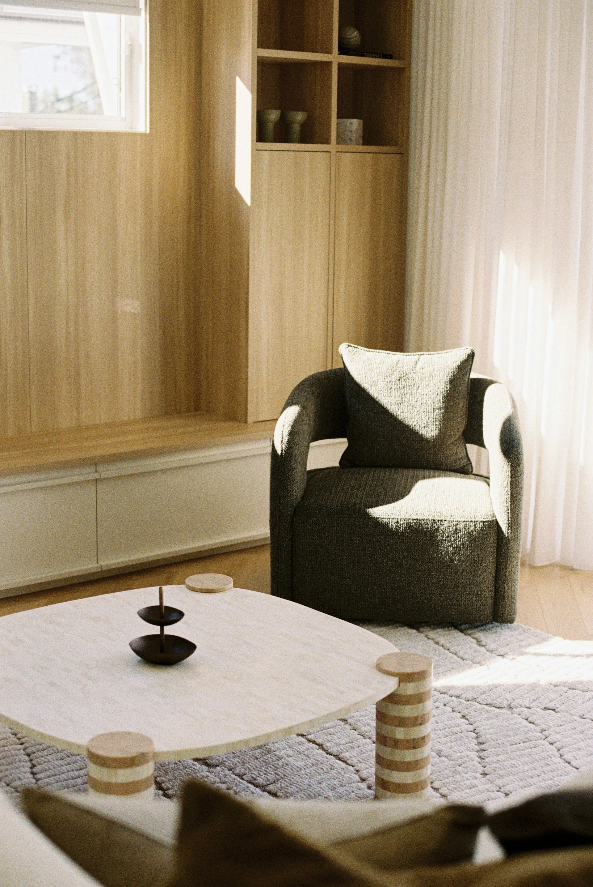 Sala moderna con sillón gris oscuro con cojín negro, mesa de centro de madera clara, estantería de madera y cortinas blancas que dejan pasar la luz.
