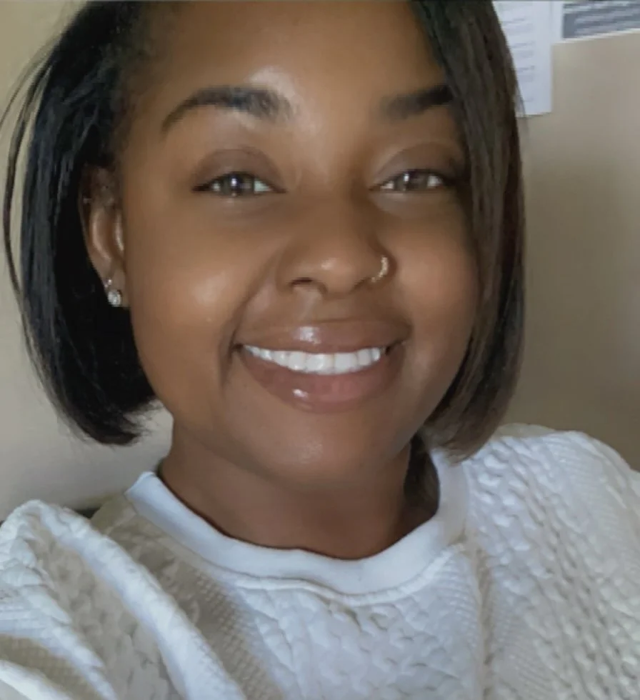 A smiling woman with dark hair, wearing small earrings and a white textured top, in an indoor setting.