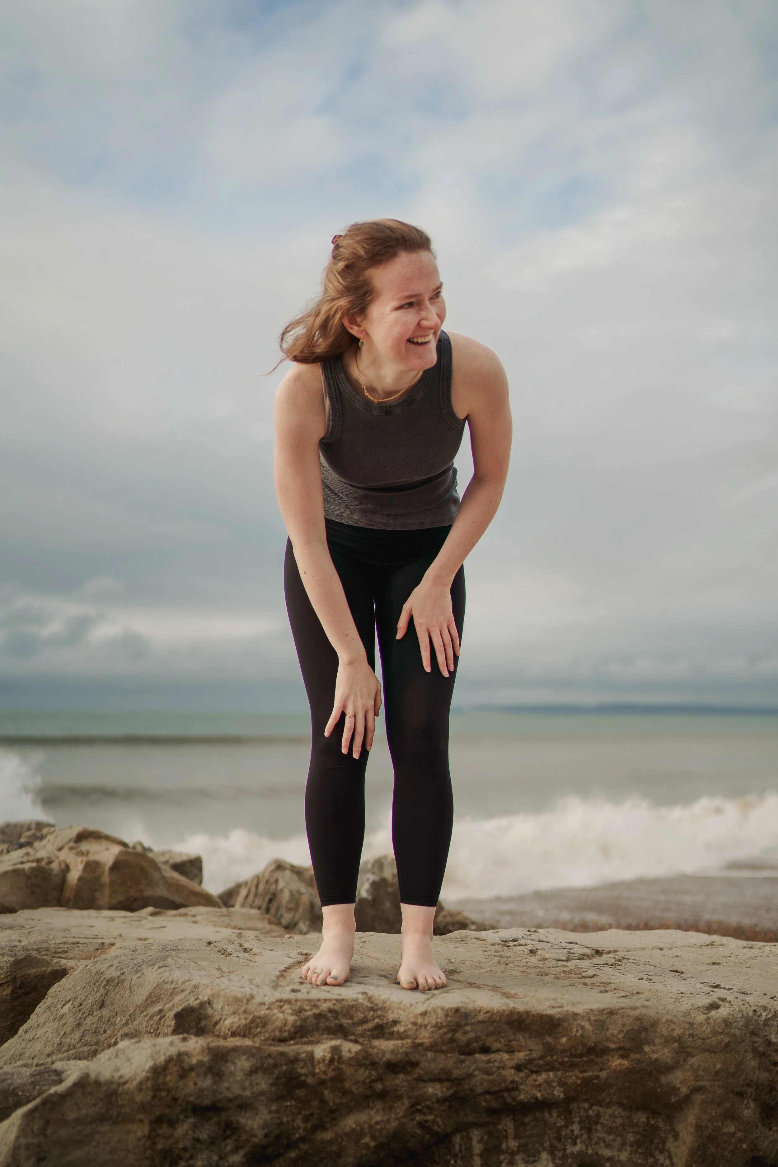A woman with curly red hair stands barefoot on large rocks at the beach, laughing and leaning forward with her hands on her knees, with waves and a cloudy sky in the background.