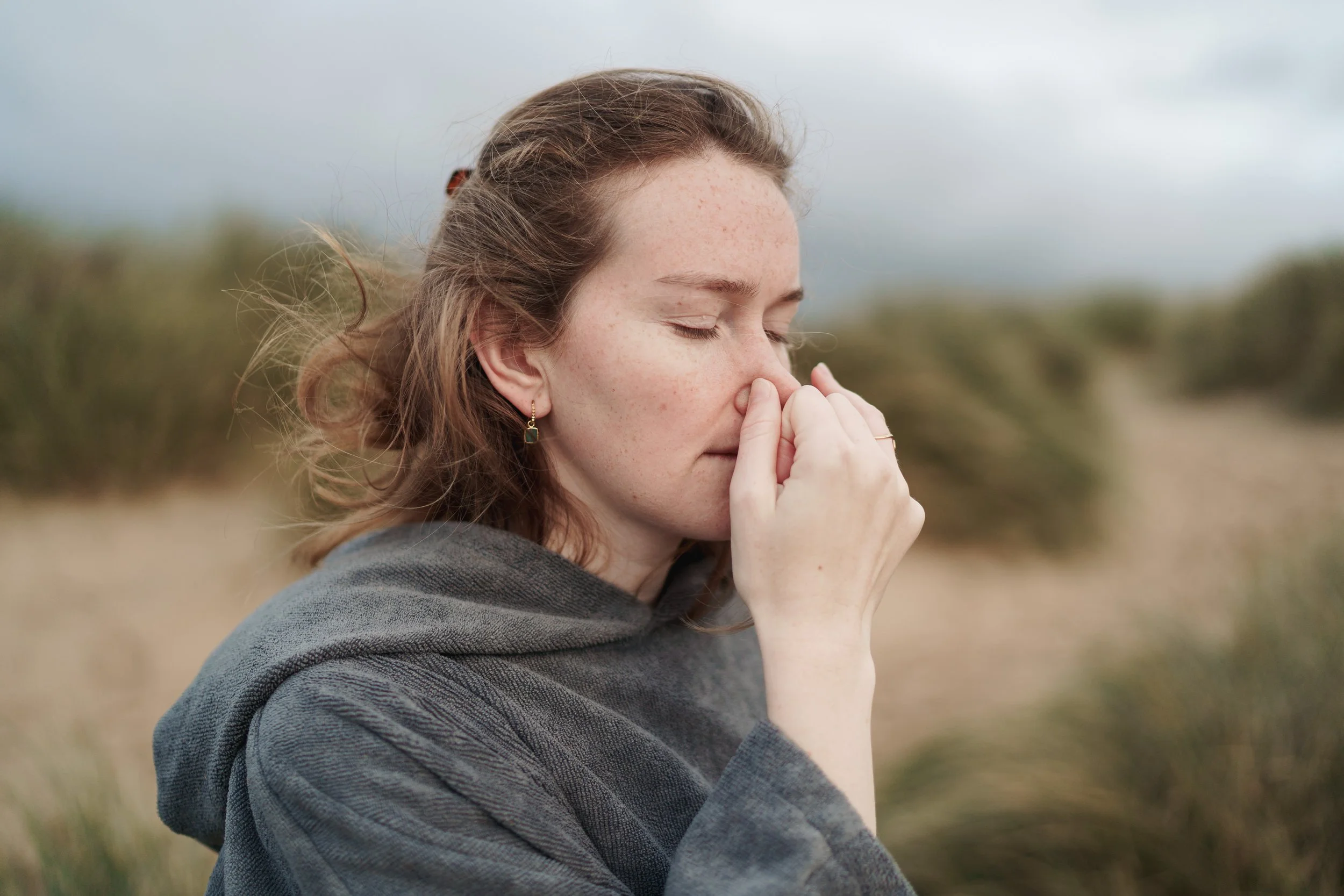 A young woman with red hair and freckles standing outdoors with her eyes closed and her hand over her nose, wearing a gray hoodie, against a blurred natural background.