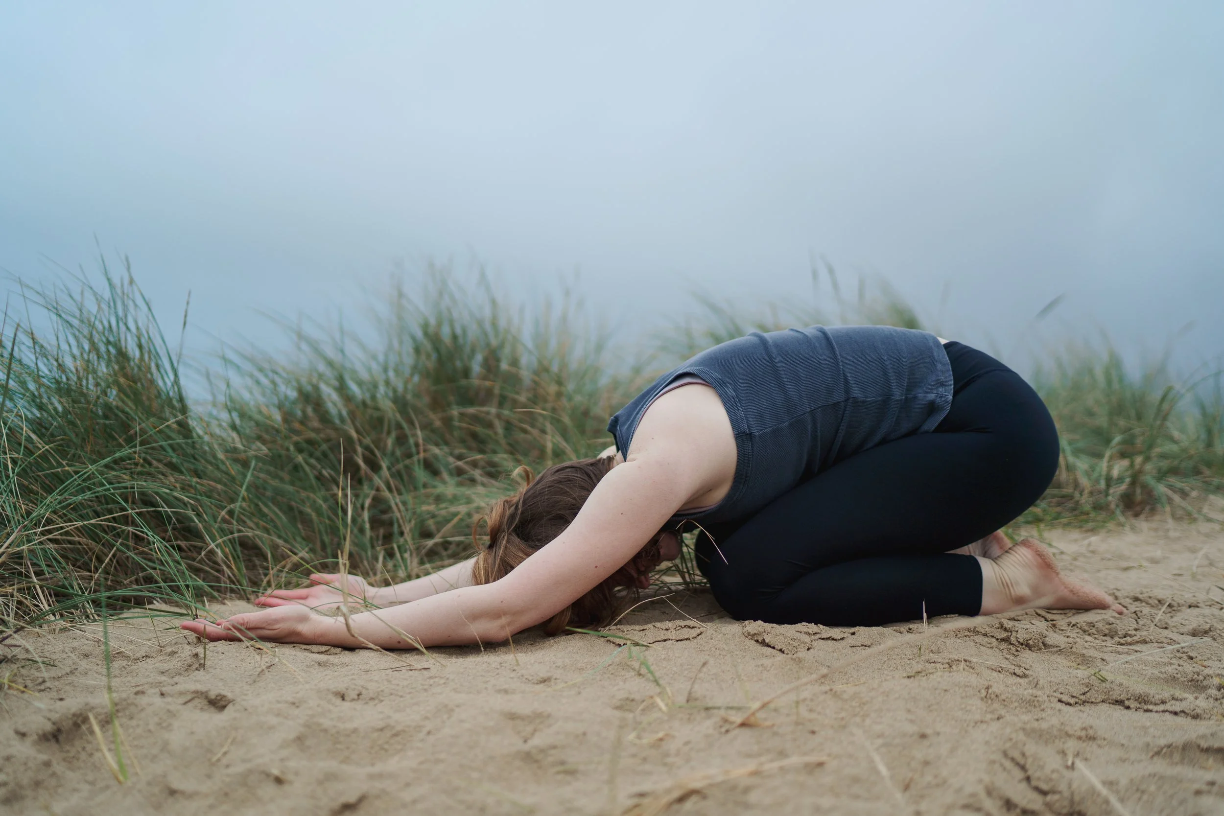 A woman practicing yoga in a child's pose on a sandy beach with tall grass and foggy sky in the background.