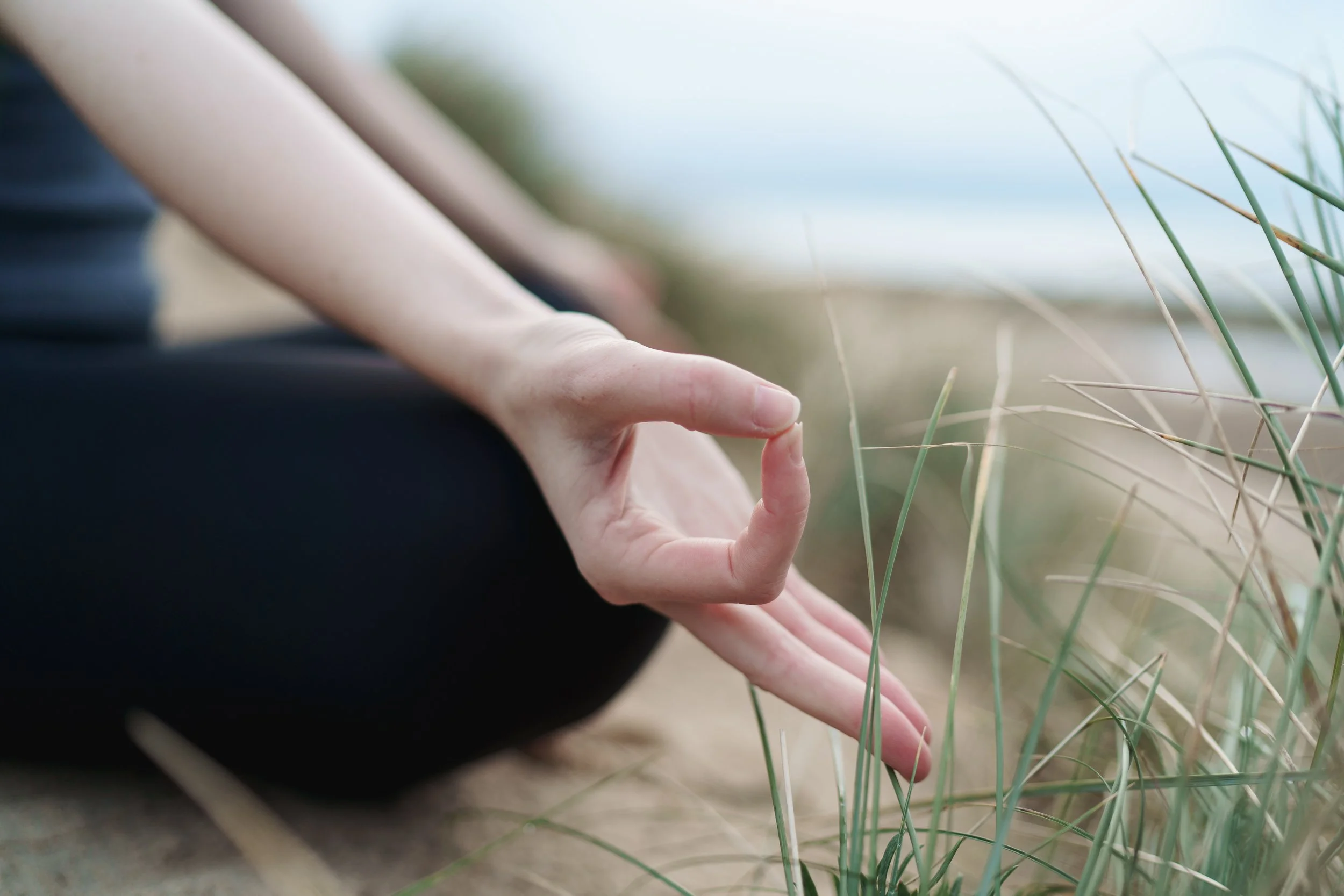 Person in a seated meditation pose on the beach, with their hand resting on their knee and fingers forming a mudra, surrounded by beach grass.