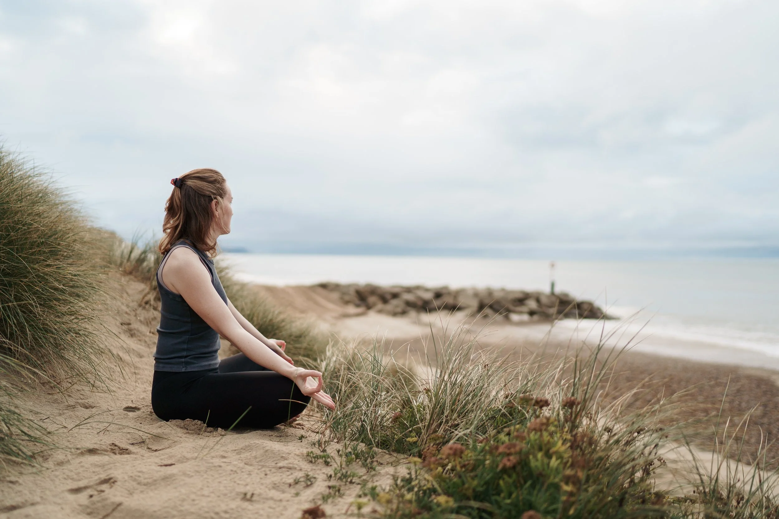 Woman sitting in a meditative pose on a sandy beach, facing the ocean, surrounded by dunes and ocean vegetation.