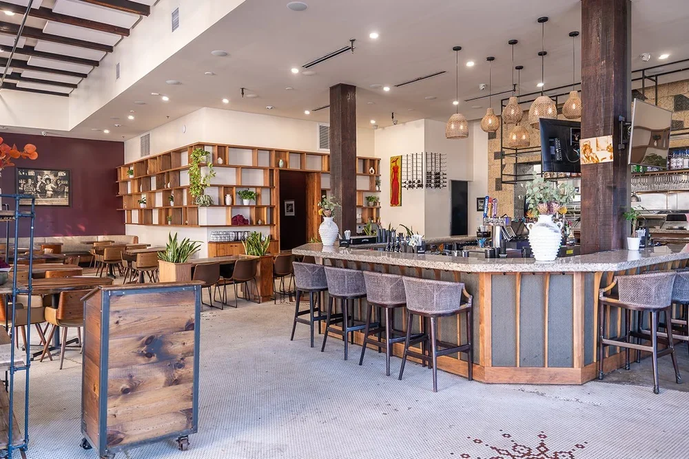 Interior of a modern restaurant or cafe with a curved bar counter, hanging pendant lights, bar stools, and wooden shelving with decorative items.