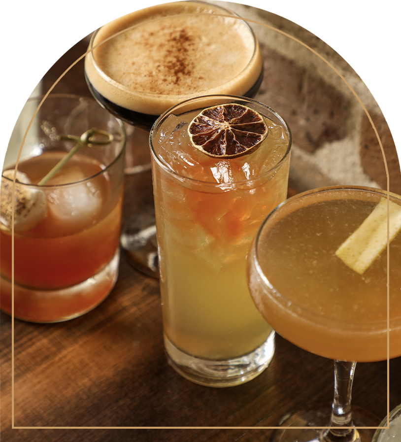 Four cocktails on a wooden table, including a margarita, a whiskey sour, a tiki drink with a cherry and pineapples, and a dark beer.
