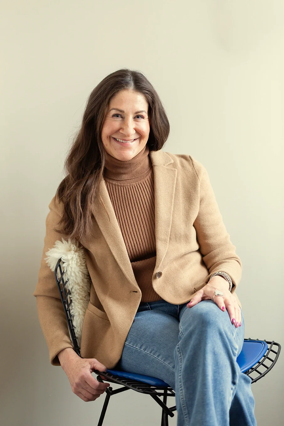 Dana Marcus, therapist & coach specializing in ADHD, in her Philadelphia office, sitting on a black chair with a blue cushion, wearing a beige blazer over a brown turtleneck, with a beige & black watch.