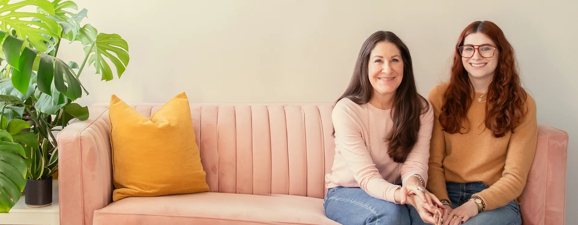 Two women sitting on a pink sofa, smiling, with a large green potted plant on the left and a yellow pillow on the sofa.