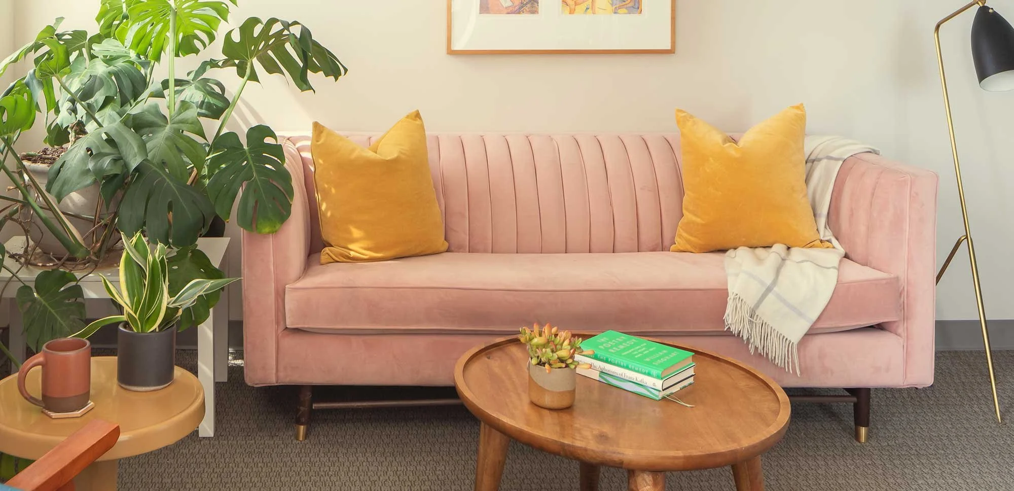 Green Fig's therapy & coaching office in Philadelphia, with a cozy pink velvet sofa, yellow pillows, a cream plaid throw blanket, a large green monstera plant, a wooden coffee table with a potted succulent & books, and framed artwork on the wall.