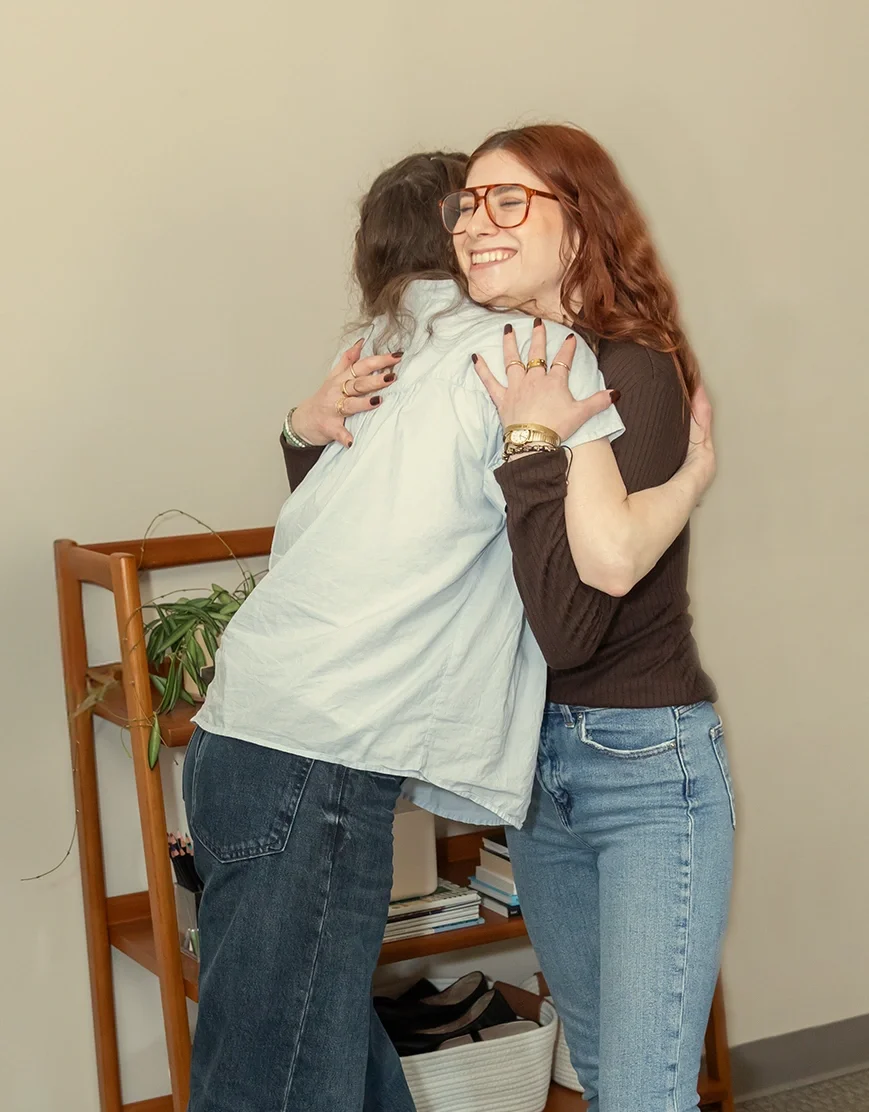 Two women hugging and smiling, standing in a room with a bookshelf and a plant.