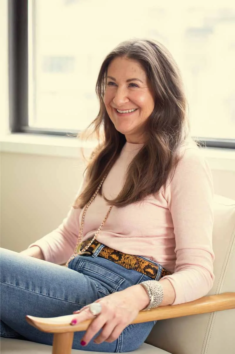 Dana Marcus, therapist & coach specializing in ADHD, sitting on a white chair near a window in her Philadelphia office, wearing a pink top, jeans, and a patterned belt.