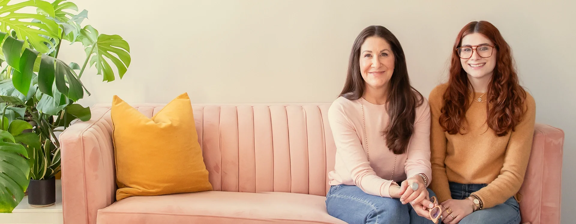 Dana Marcus, therapist & coach specializing in ADHD, and Molly Marcus, therapist & coach specializing in DBT & Decluttering, sitting together on a pink velvet sofa with yellow cushions and a large green plant to the side, smiling at the camera.
