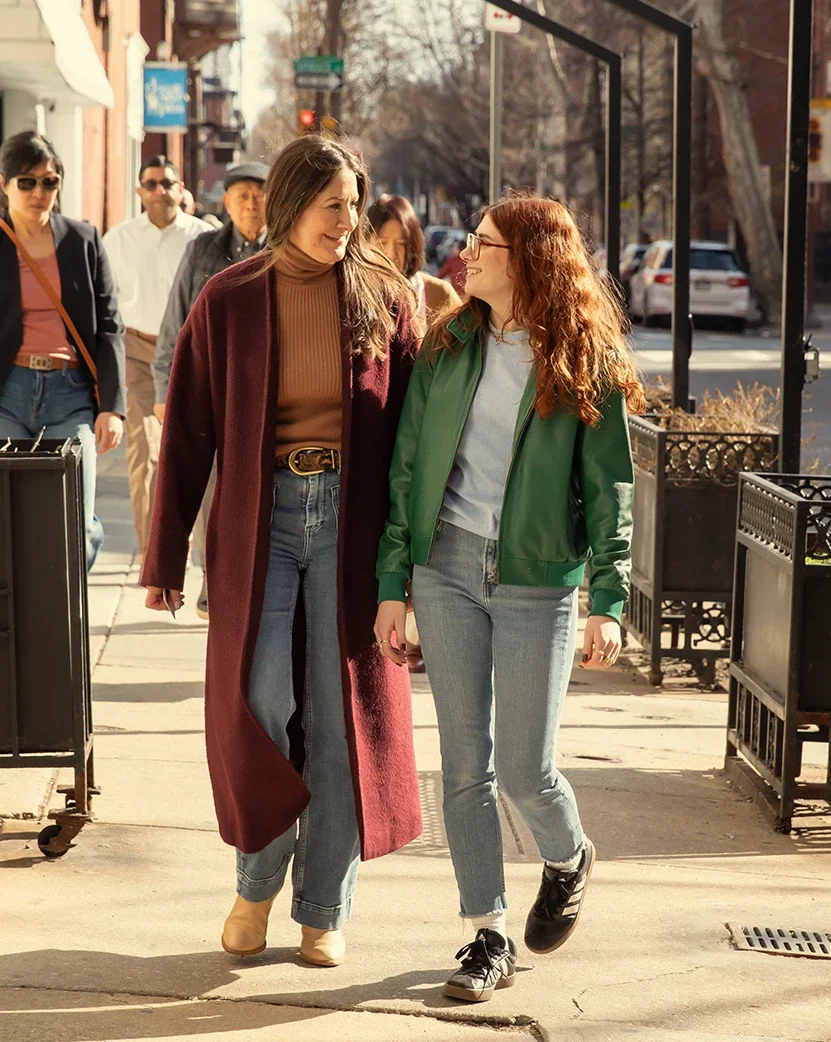 Dana Marcus, wearing a long plum coat, and Molly Marcus, wearing a green leather jacket, walking on the sidewalk outside of their therapy & coaching office in Philadelphia.