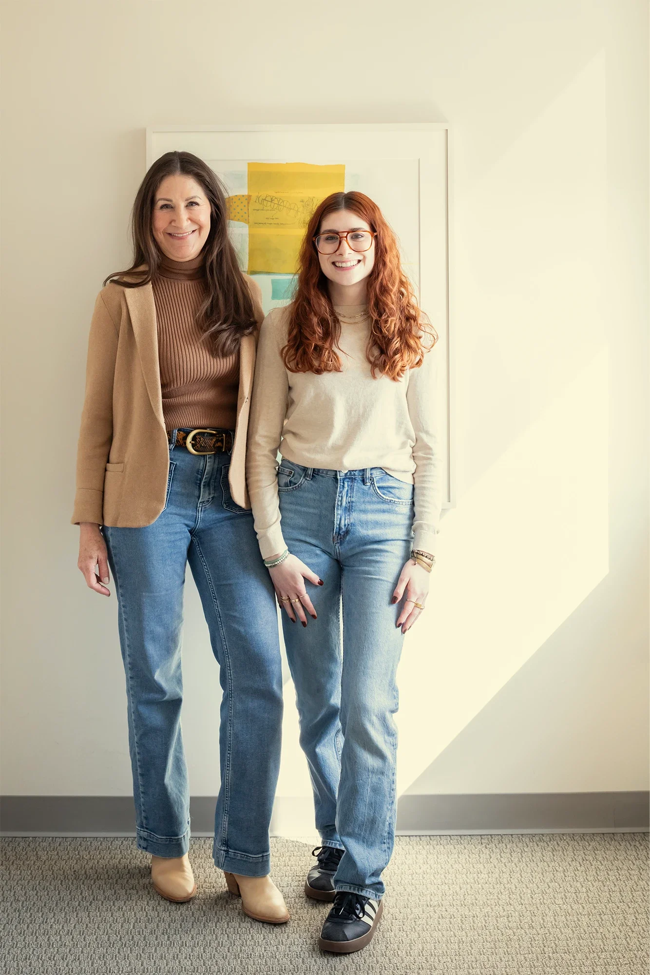 Two women standing together in an office, smiling at the camera. The woman on the left has long dark hair and is wearing a beige blazer, brown turtleneck, and jeans. The woman on the right has curly red hair, glasses, and is wearing a cream sweater and jeans. There is a framed artwork behind them on the wall.