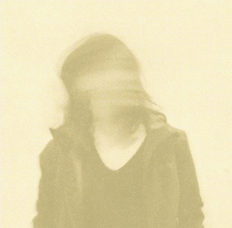 A blurry faded, yellowed image of a person with shoulder-length hair wearing a dark jacket and shirt, standing against a plain background.