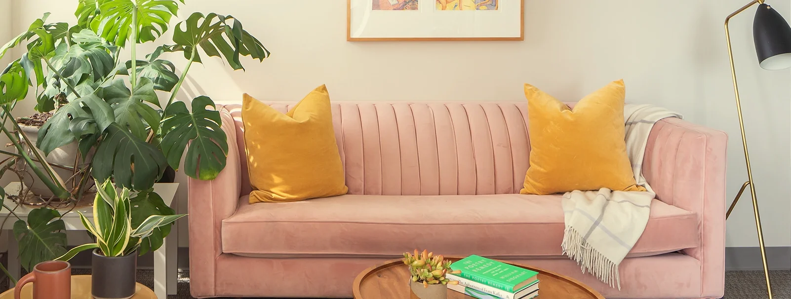Green Fig's therapy & coaching office in Philadelphia, with a cozy pink velvet sofa, yellow pillows, a cream plaid throw blanket, a large green monstera plant, a wooden coffee table with a potted succulent & books, and framed artwork on the wall.