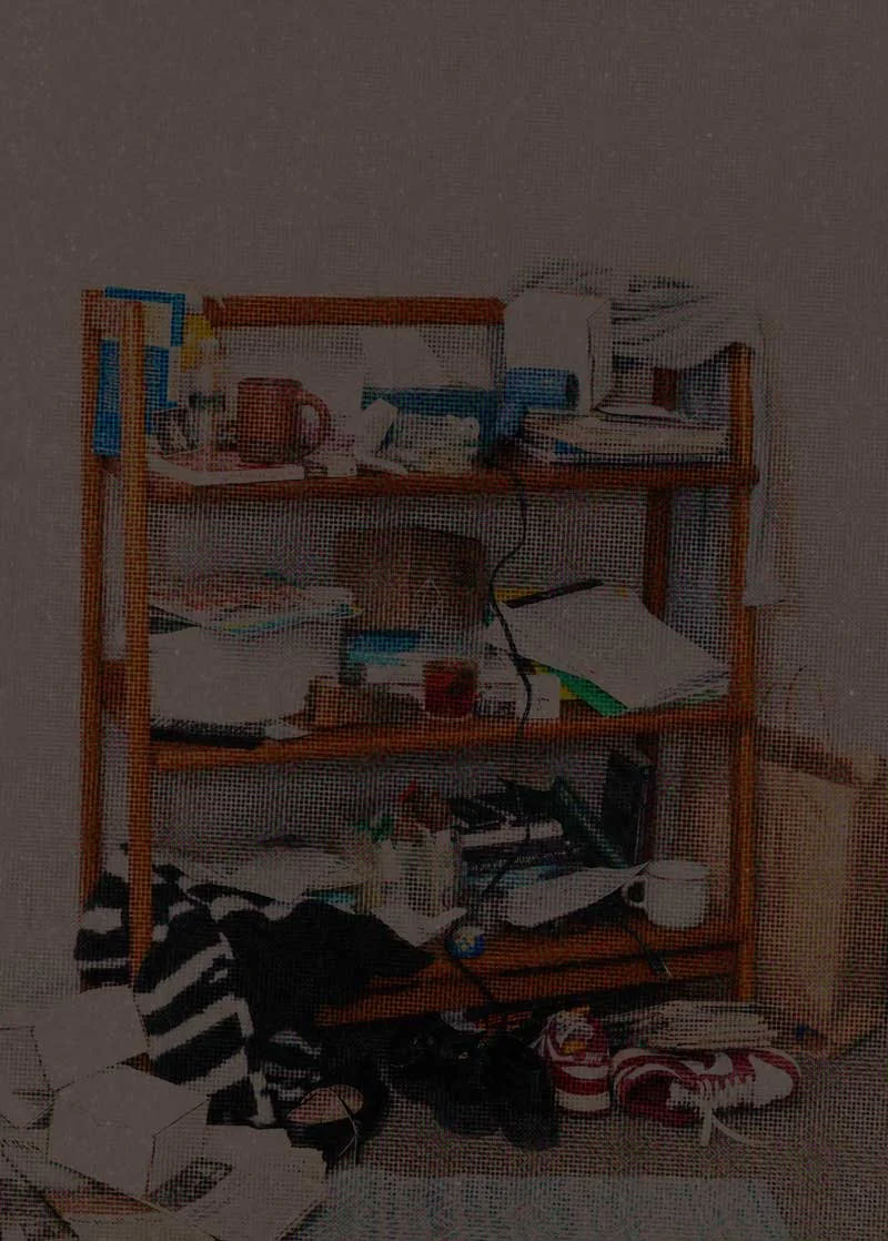 A  very cluttered & disorganized wooden shelf with various items including books, papers, and electronic devices, situated against a wall.
