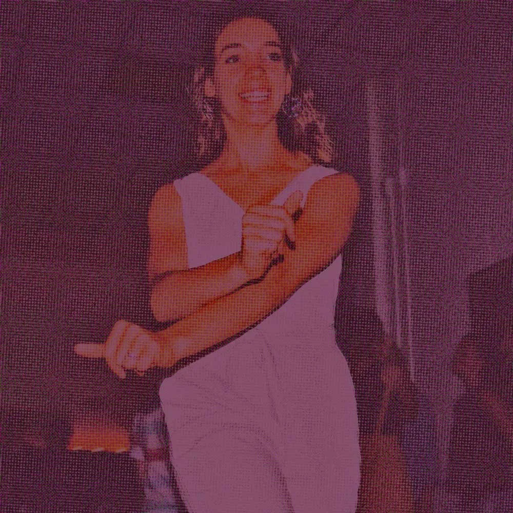 A woman smiling, wearing a white dress, standing indoors with her hands clasped in front of her.